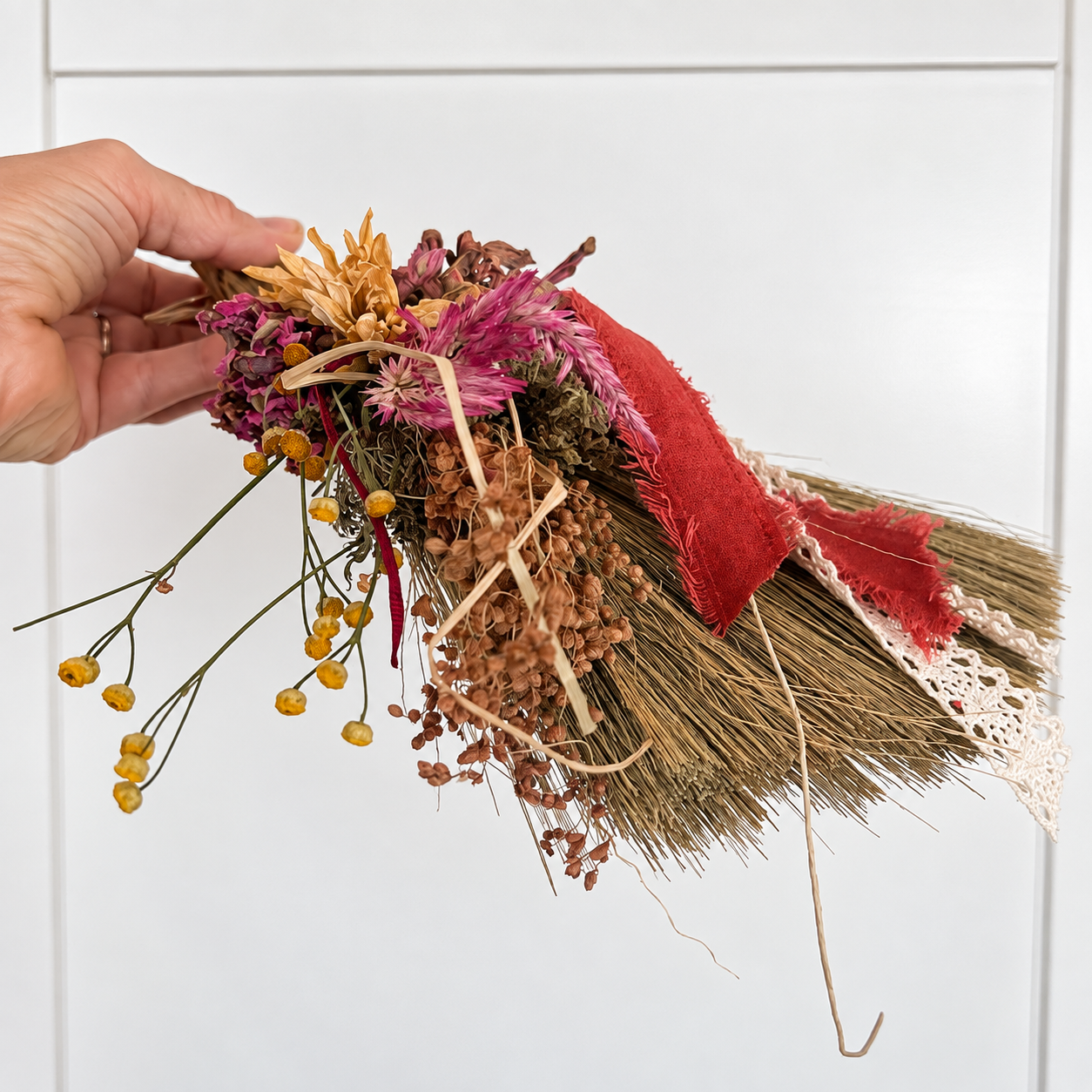 Close-up of dried flowers, seed stems, red ribbon and lace arranged by hand on The Hearthkeeper’s Botanical Broom.