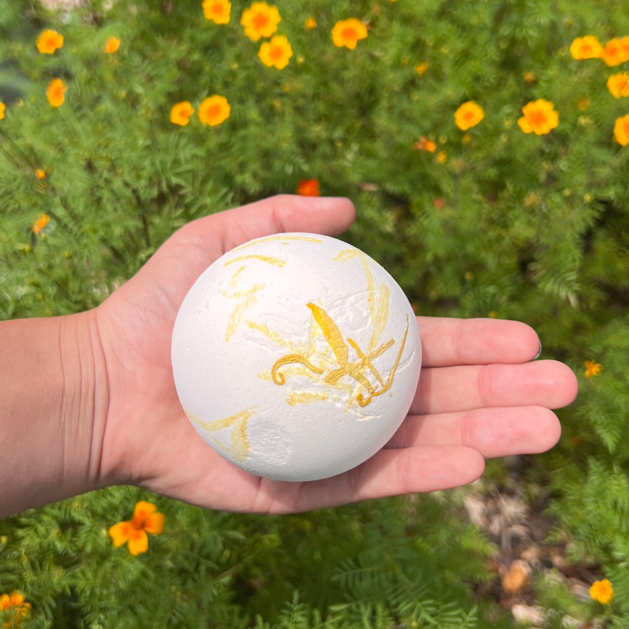 Handcrafted Honey Bath Bomb with real calendula petals resting in a child's hand in a blooming flower garden. Natural honey-scented bath bomb made with botanical ingredients for kids and gift giving by Sow the Magic®.
