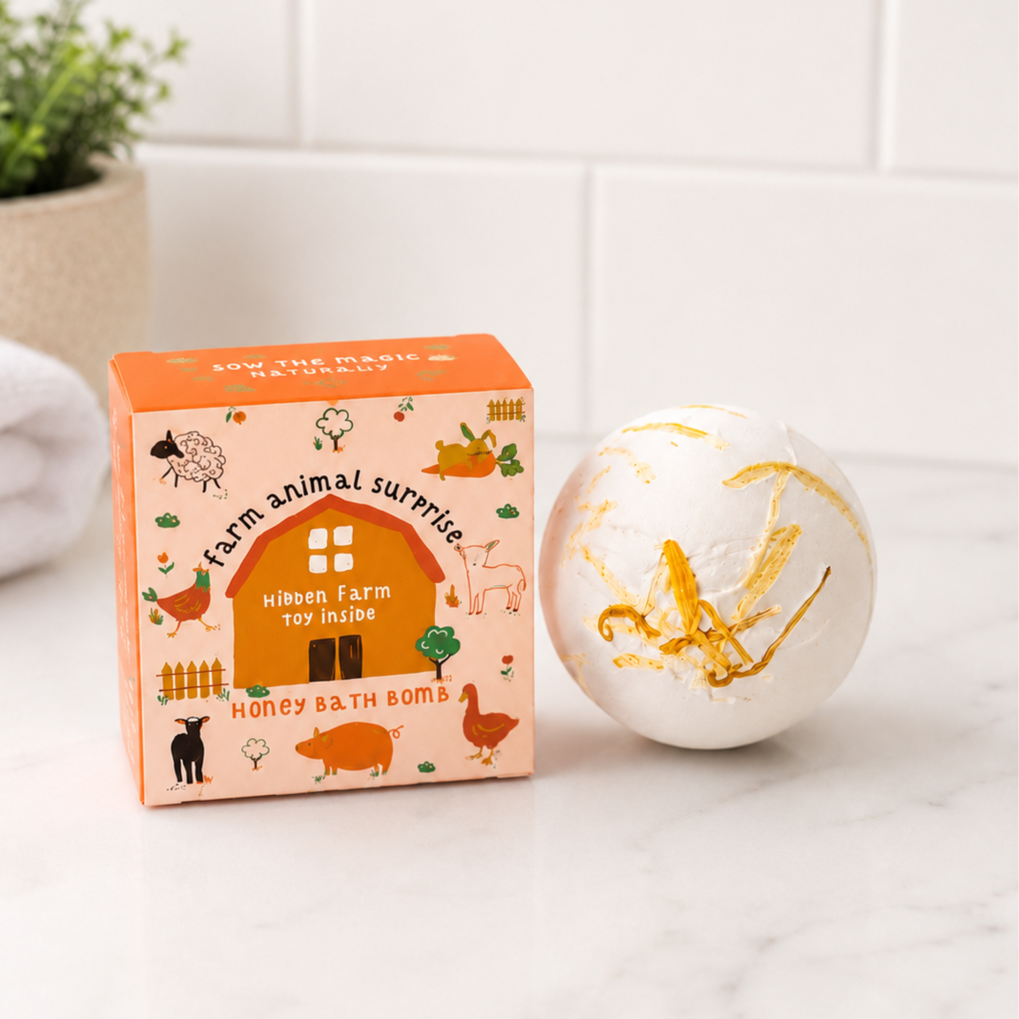 Sow the Magic® Honey Bath Bomb with a hidden farm animal toy inside displayed beside its gift box on a white marble bathroom countertop. Honey-scented kids surprise bath bomb handcrafted with natural ingredients and topped with real calendula petals for a fun, fizzy bath time experience.