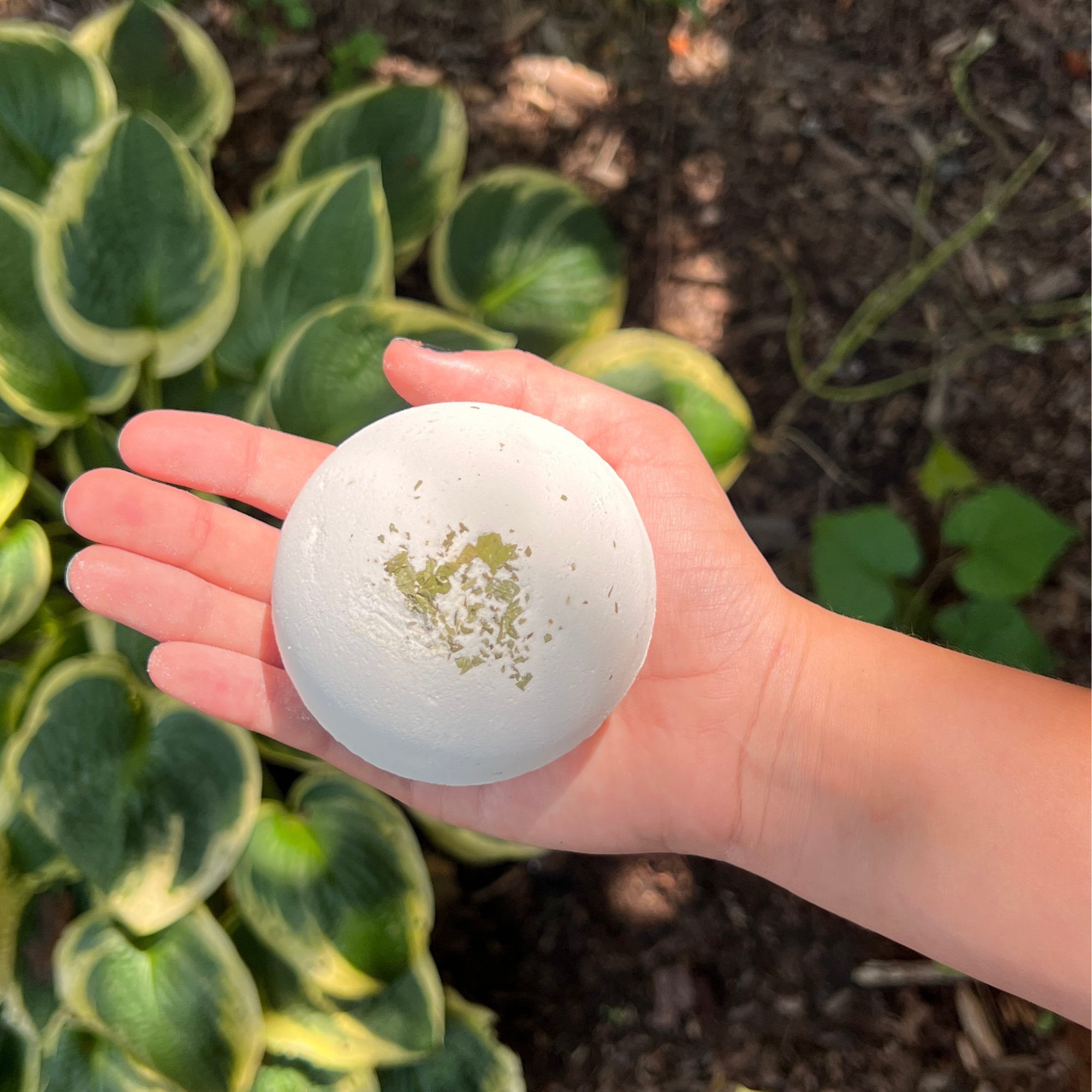 Child holding the Sow the Magic® Lemon Bath Bomb topped with real peppermint leaves in a garden setting. Handmade kids bath bomb with a hidden bug toy inside, crafted with shea butter, cocoa butter, and botanical extracts for fun, fizzy bath time adventures.
