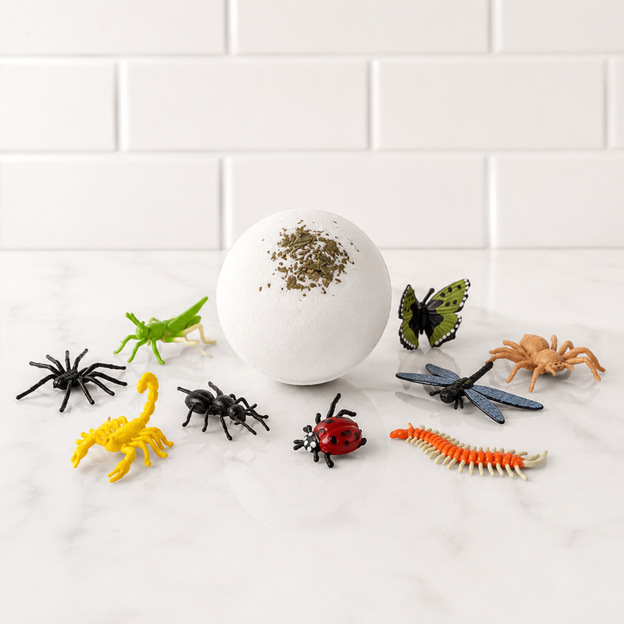 Handmade Lemon Bath Bomb topped with real peppermint leaves surrounded by collectible bug toys including a butterfly, ladybug, dragonfly, grasshopper, spiders, scorpion, beetle, centipede, and spider on a white marble tile background. Hidden bug toy bath bomb for kids by Sow the Magic®, made with shea butter and cocoa butter.