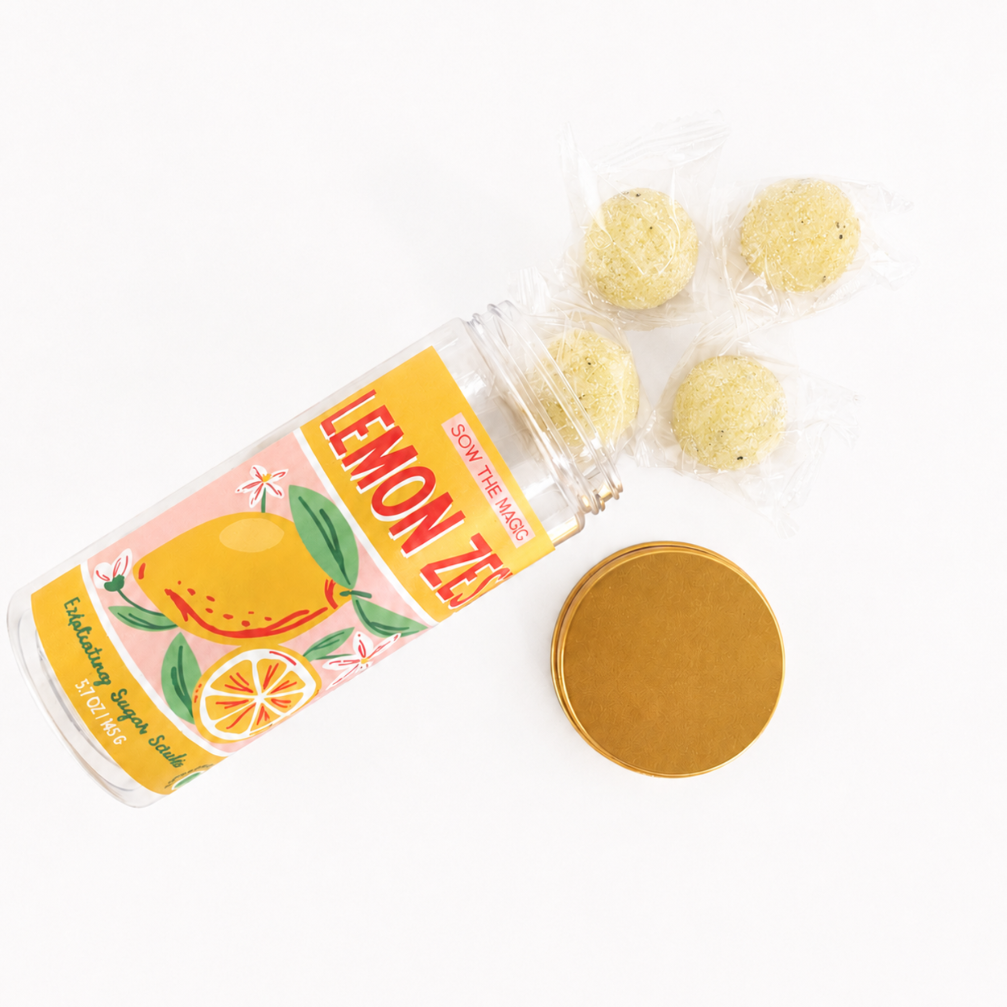 Open jar of Lemon Zest exfoliating sugar scrub cubes with gold lid and individually wrapped sugar scrub cubes arranged on a clean white background.