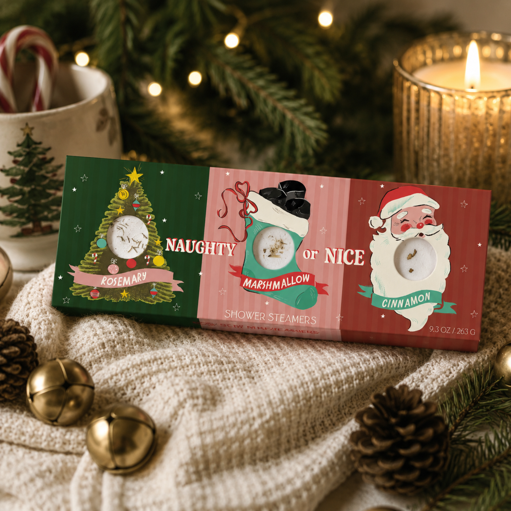Festive lifestyle photo of the Sow the Magic Naughty or Nice Christmas Shower Steamer Gift Set displayed on a cozy knit blanket with evergreen branches, pinecones, jingle bells, a glowing candle and holiday lights. The gift box includes three aromatherapy shower steamers in Rosemary, Marshmallow and Cinnamon, making it a whimsical Christmas self-care gift.