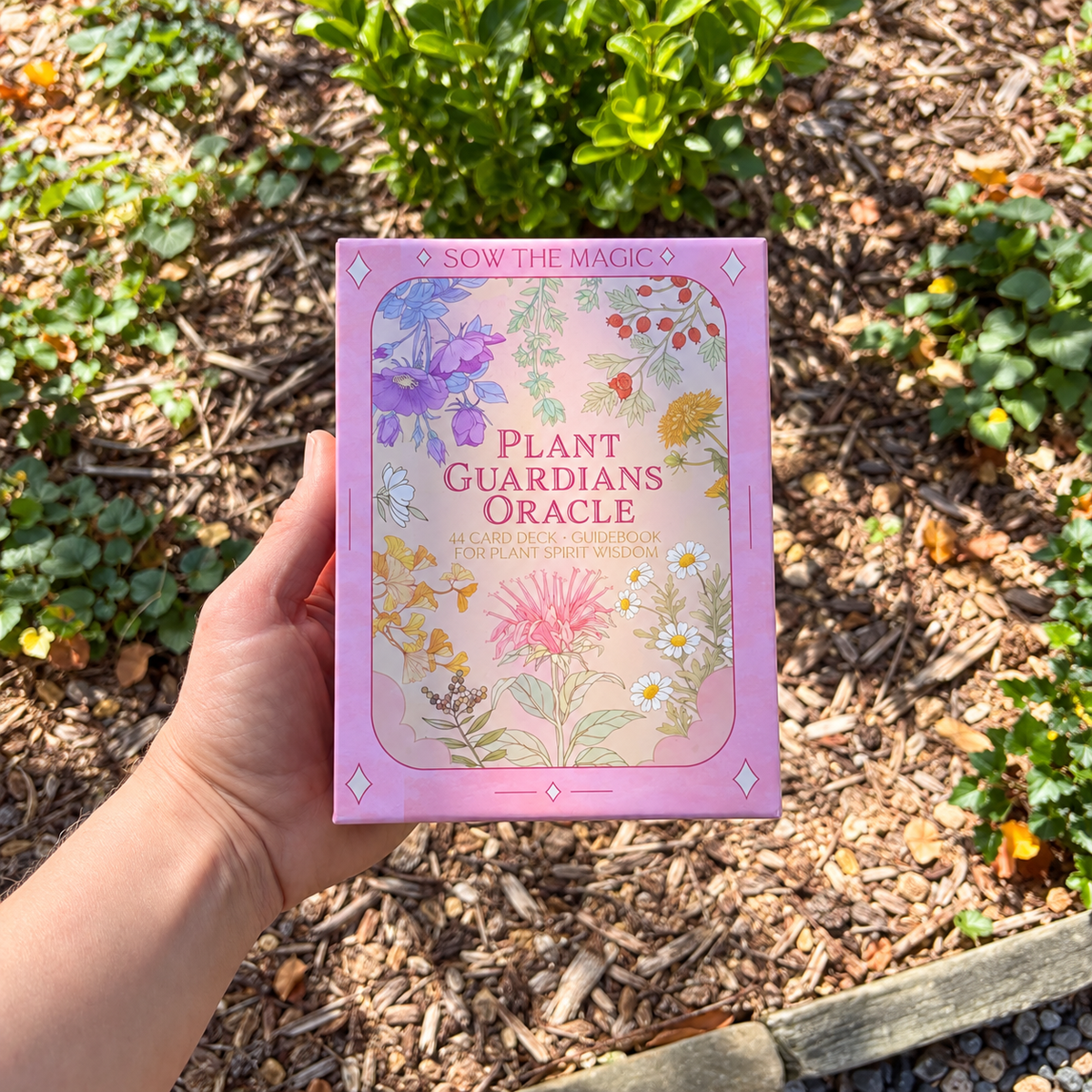 Hand holding the Sow the Magic Plant Guardians Oracle 44-card deck in its pink floral keepsake box outdoors, surrounded by natural garden greenery.