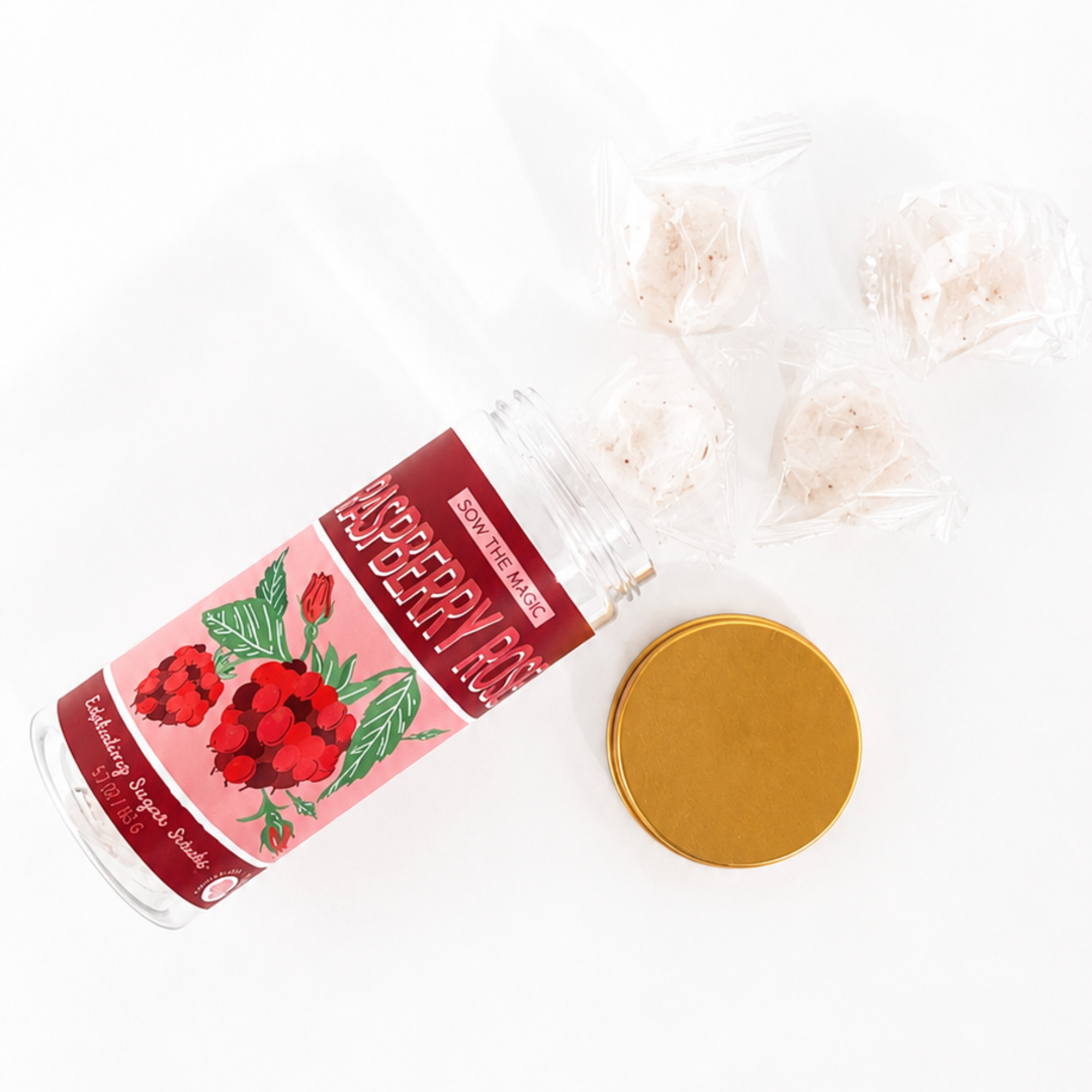 Open Raspberry Rose sugar scrub jar with gold lid and individually wrapped exfoliating sugar scrub cubes arranged on a bright white background.