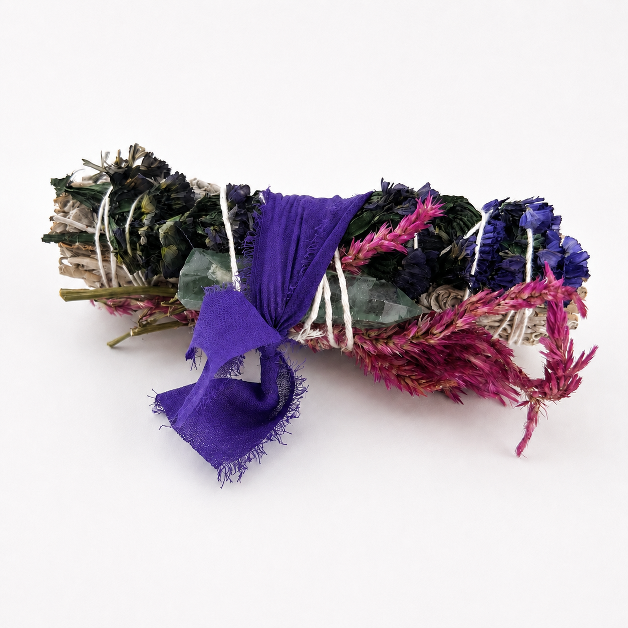 Side view of a white sage cleansing bundle wrapped with celosia flowers, blue botanicals, fluorite and purple ribbon