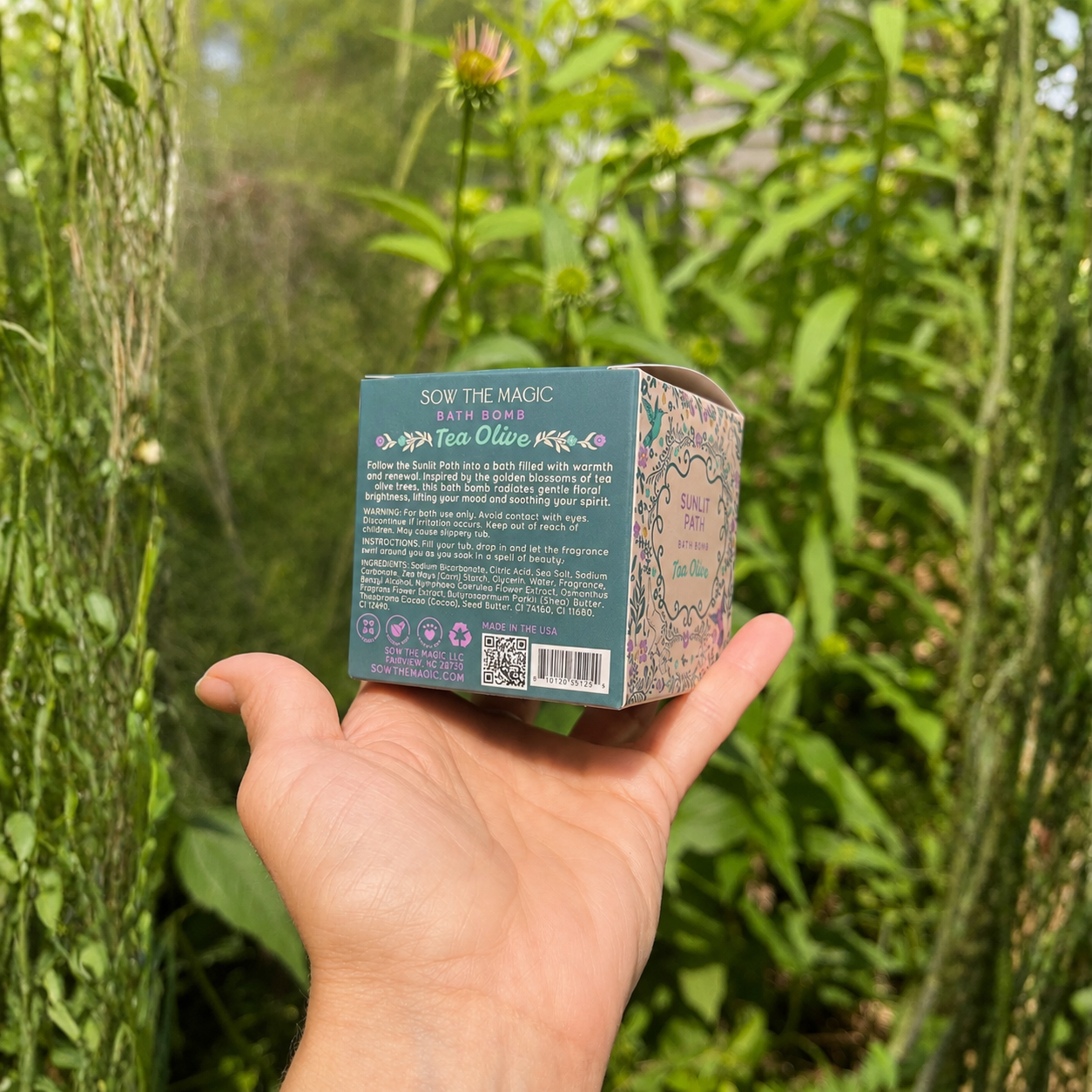 Hand holding the back of the Sow the Magic Sunlit Path Tea Olive bath bomb box in a lush summer garden with soft natural light, showcasing the product details against blooming greenery and a dreamy botanical background.