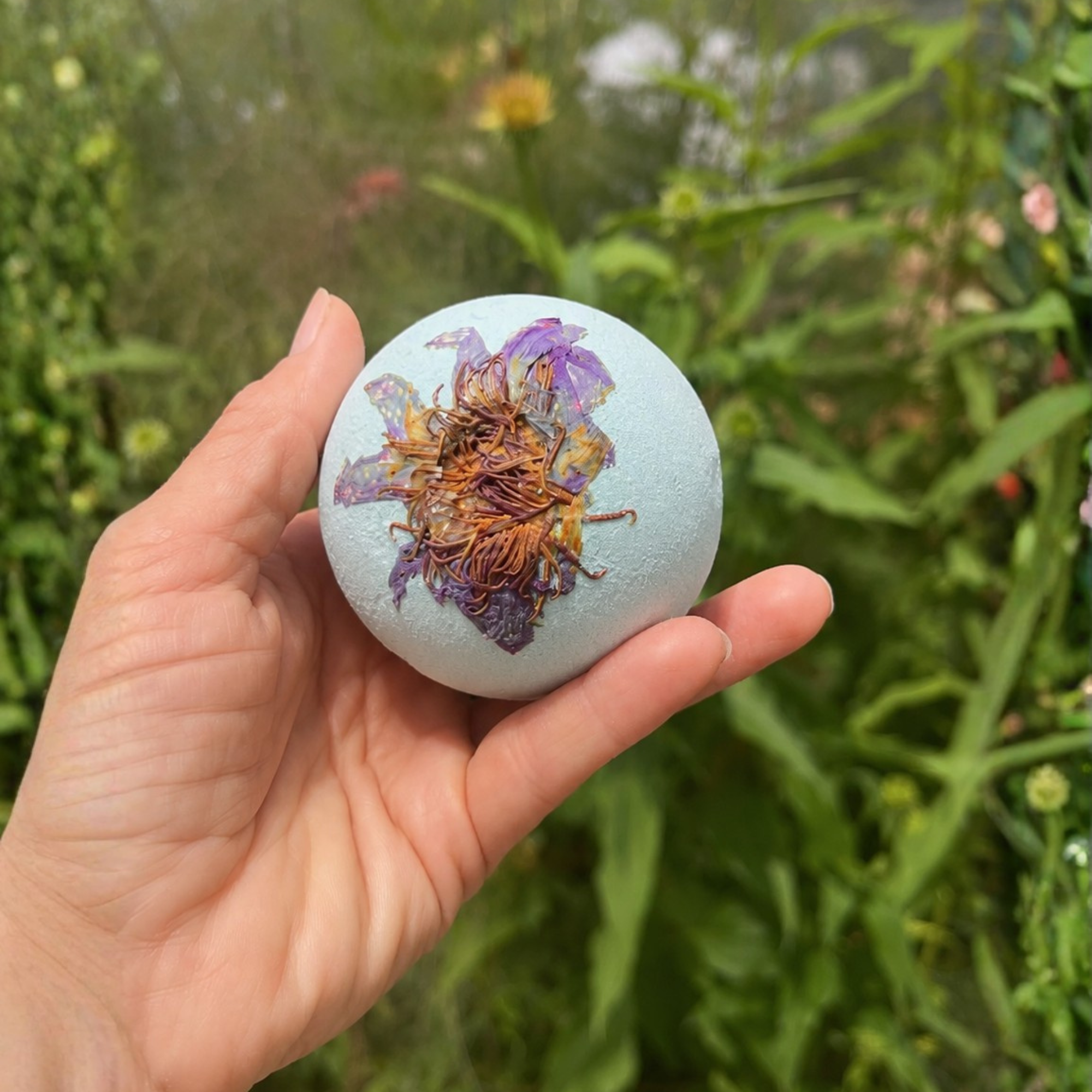 Hand holding a Sunlit Path Tea Olive botanical bath bomb topped with a dried lotus flower in a lush garden setting with soft natural lighting and blurred greenery.