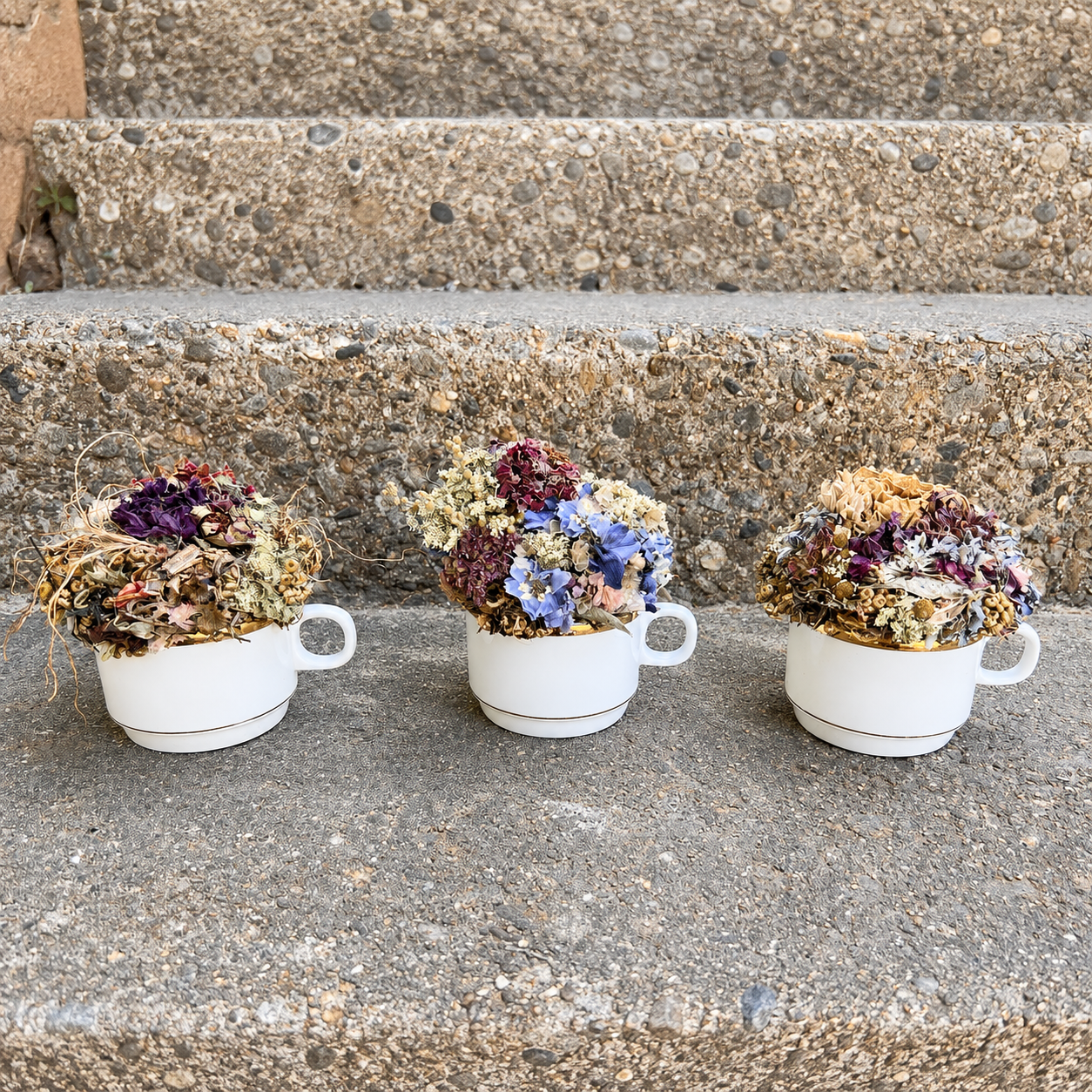 Three handmade dried flower teacup arrangements displayed together on outdoor stone steps.