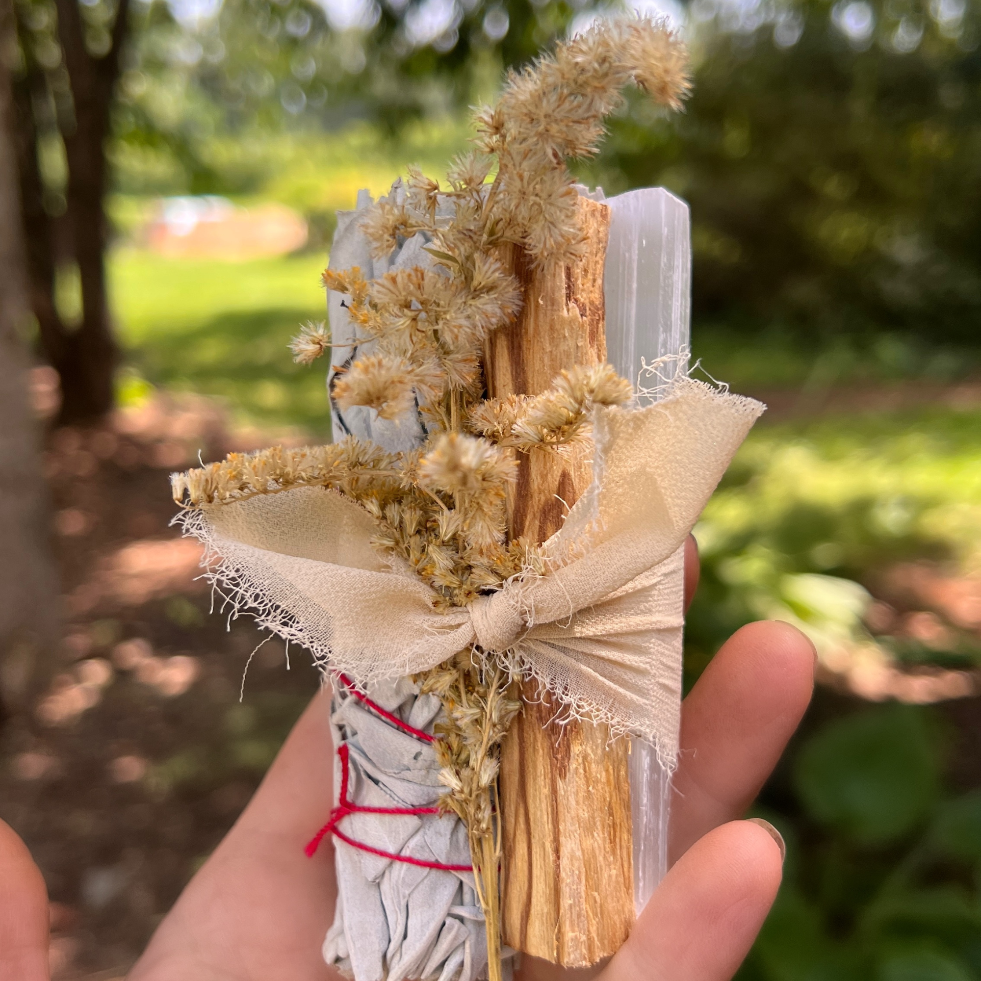 Hand holding a handcrafted white sage smudge bundle with palo santo, selenite crystal and dried goldenrod outdoors in a lush garden setting.