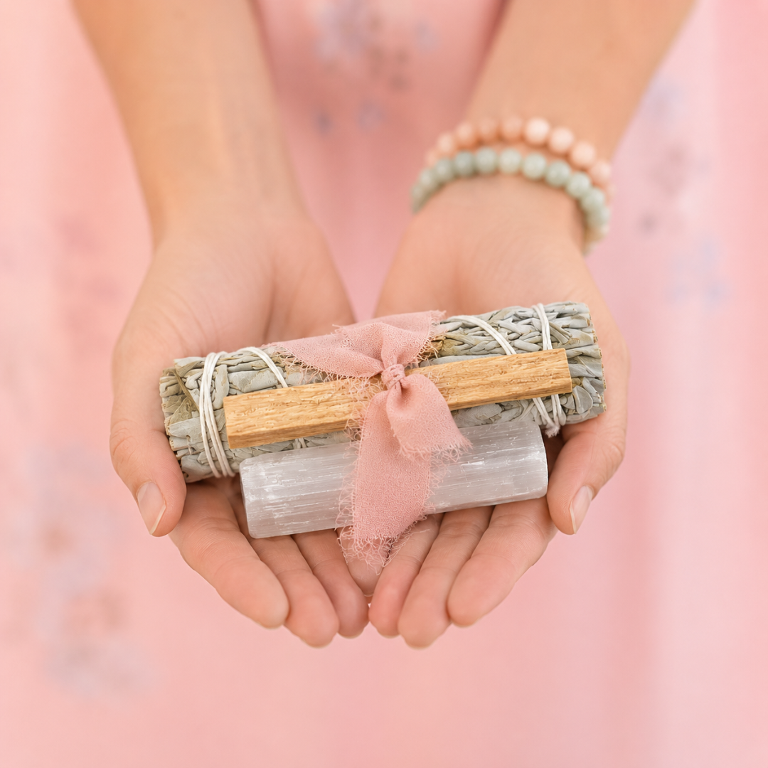 Hands gently holding a handcrafted White Sage Protection Ritual Bundle featuring White Sage, a Palo Santo stick, a genuine Selenite cleansing wand and a soft blush ribbon against a pastel pink background. Handmade spiritual gift by Sow the Magic®.