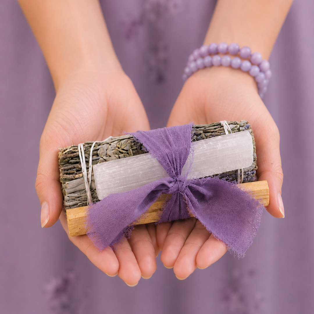 Hands gently holding a handcrafted White Sage Protection Ritual Bundle featuring White Sage, a genuine Selenite cleansing wand, Palo Santo stick and a soft lavender ribbon against a muted lavender background. Lavender gemstone bracelets add a calming touch to this handmade spiritual gift by Sow the Magic