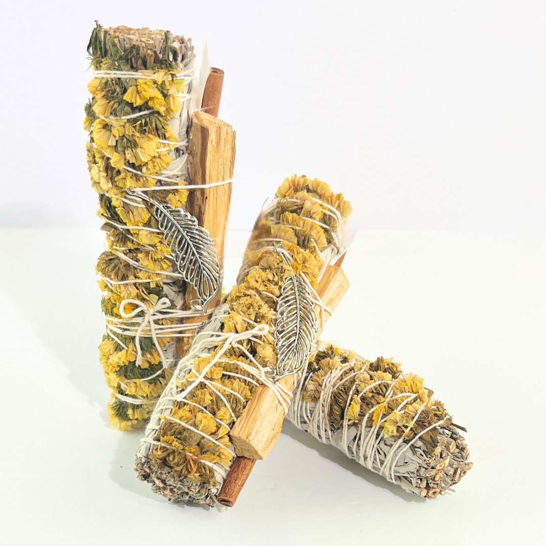 Handcrafted White Sage smudge stick wrapped with Palo Santo, yellow sinuata flowers and a silver feather charm. Handmade botanical ritual bundle on a bright white background by Sow the Magic®.