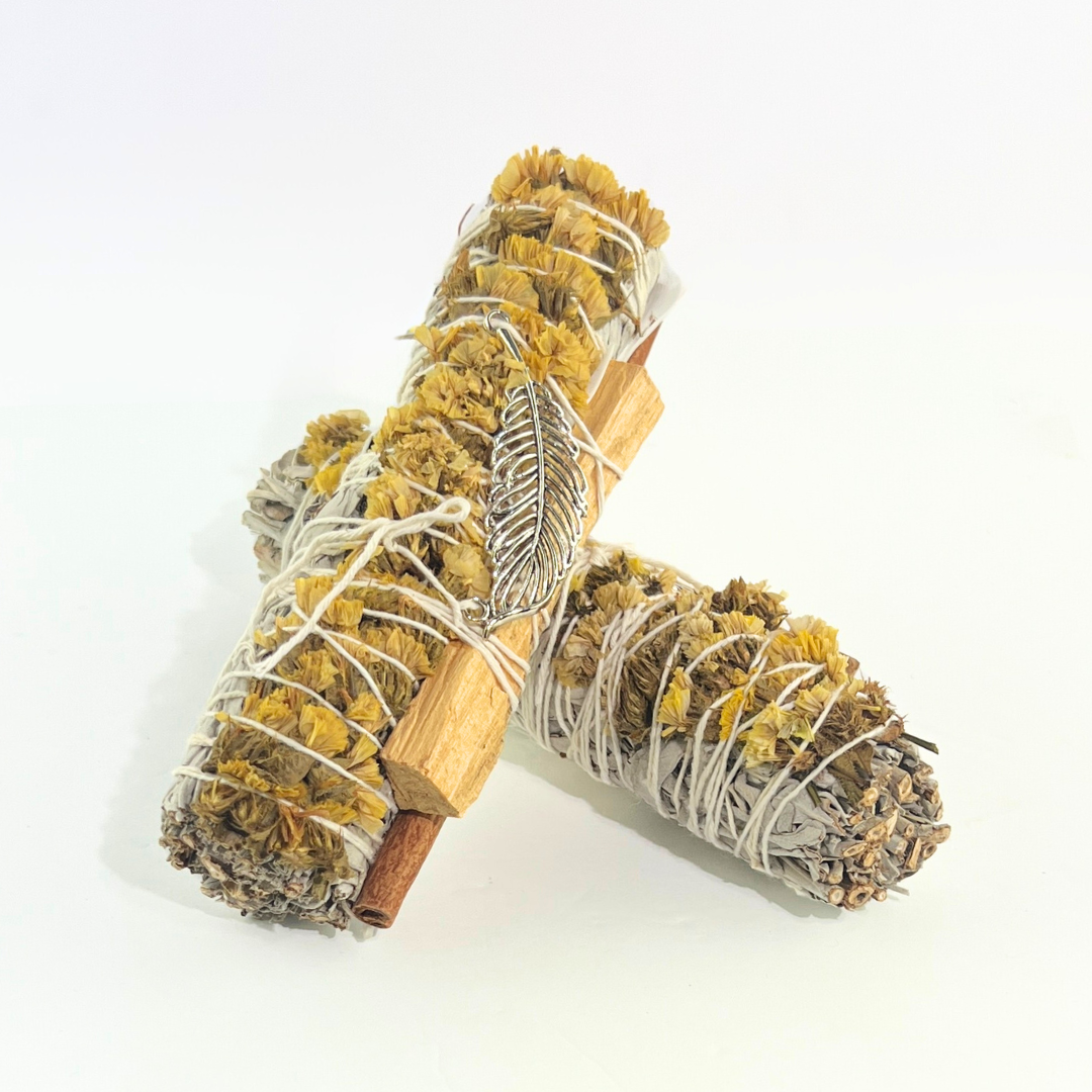 Handcrafted White Sage smudge stick wrapped with Palo Santo, yellow sinuata flowers and a silver feather charm. Handmade botanical ritual bundle on a bright white background by Sow the Magic®.
