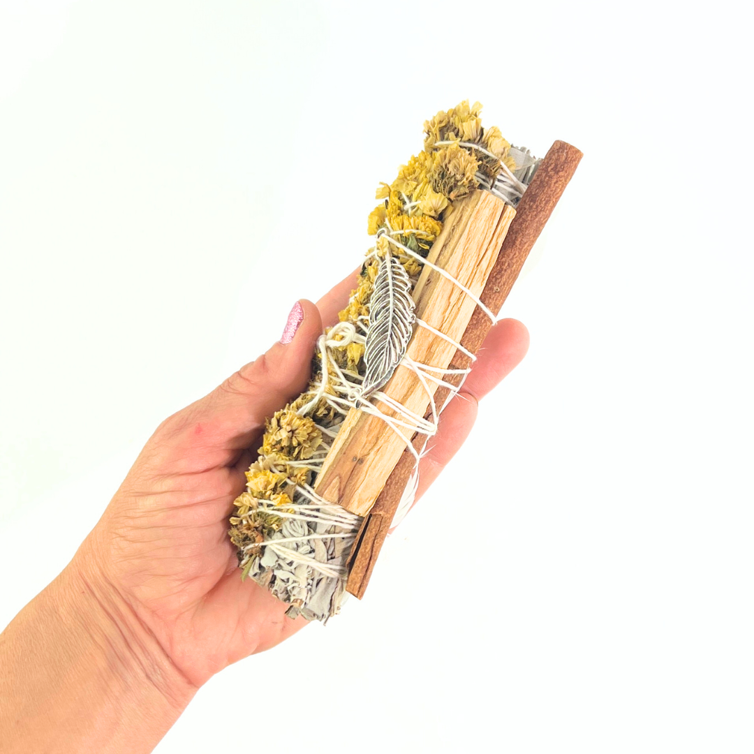 Handcrafted White Sage smudge stick wrapped with Palo Santo, yellow sinuata flowers and a silver feather charm. Handmade botanical ritual bundle on a bright white background by Sow the Magic®.