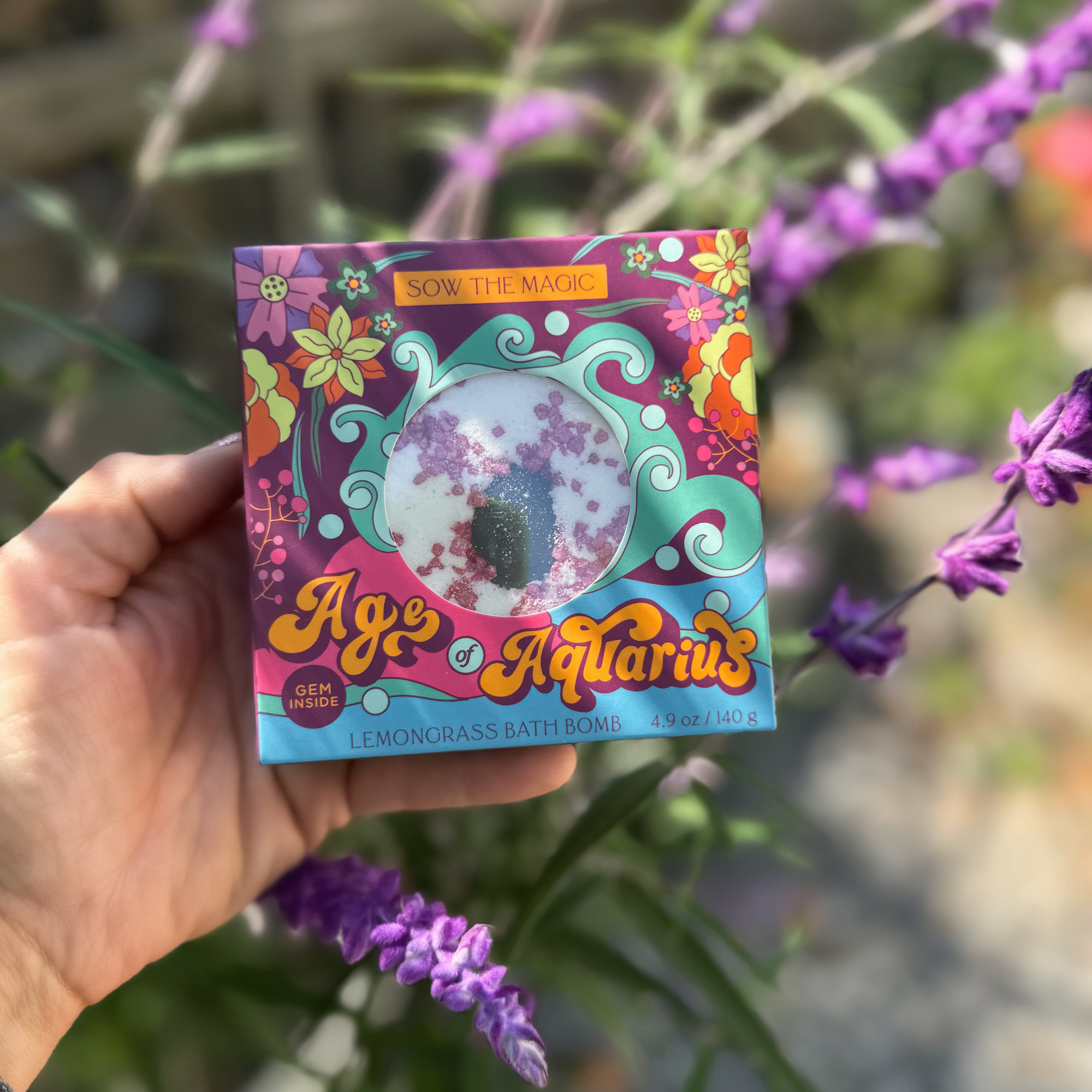 Hand holding Age of Aquarius Lemongrass Bath Bomb surrounded by blooming purple flowers outdoors