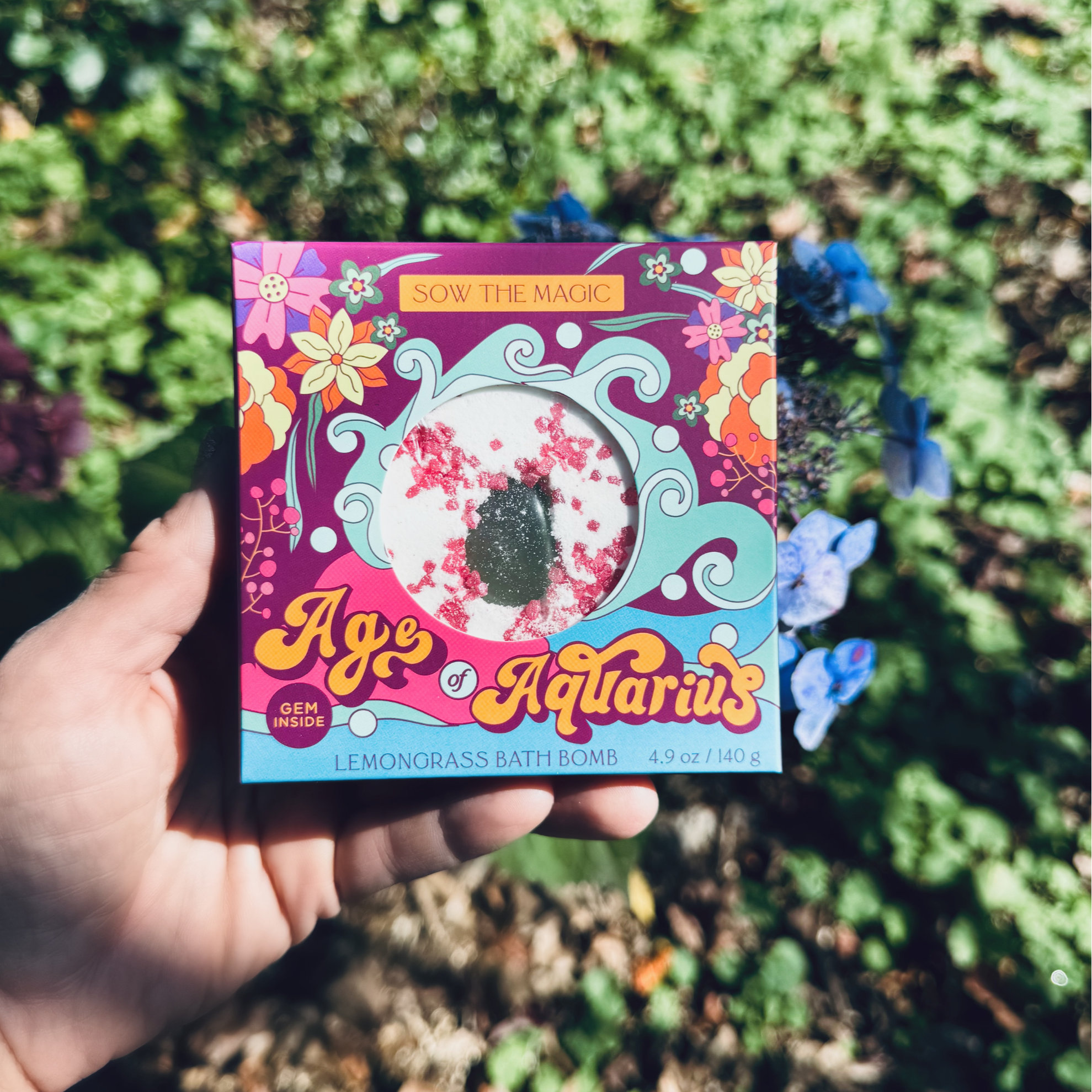 Hand holding Age of Aquarius Lemongrass Bath Bomb box outdoors surrounded by greenery