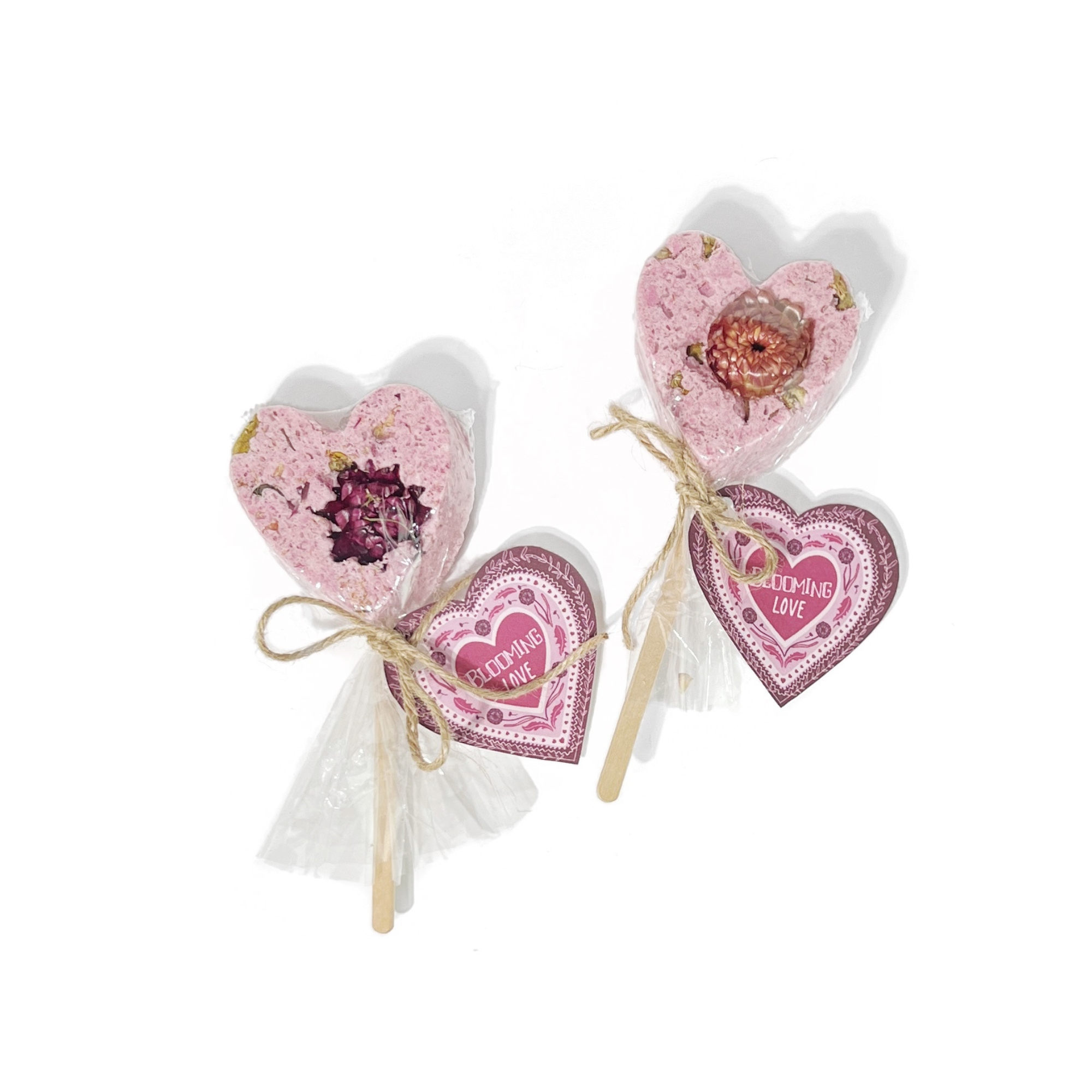 Two pink heart bath clay pops wrapped in clear packaging with “Blooming Love” tags and twine bows.