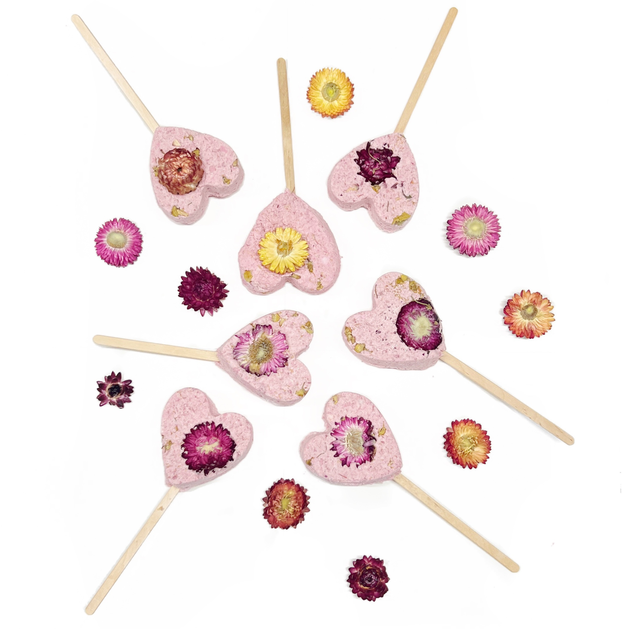 Flat lay of pink heart bath clay pops surrounded by colorful dried flowers on a white background.