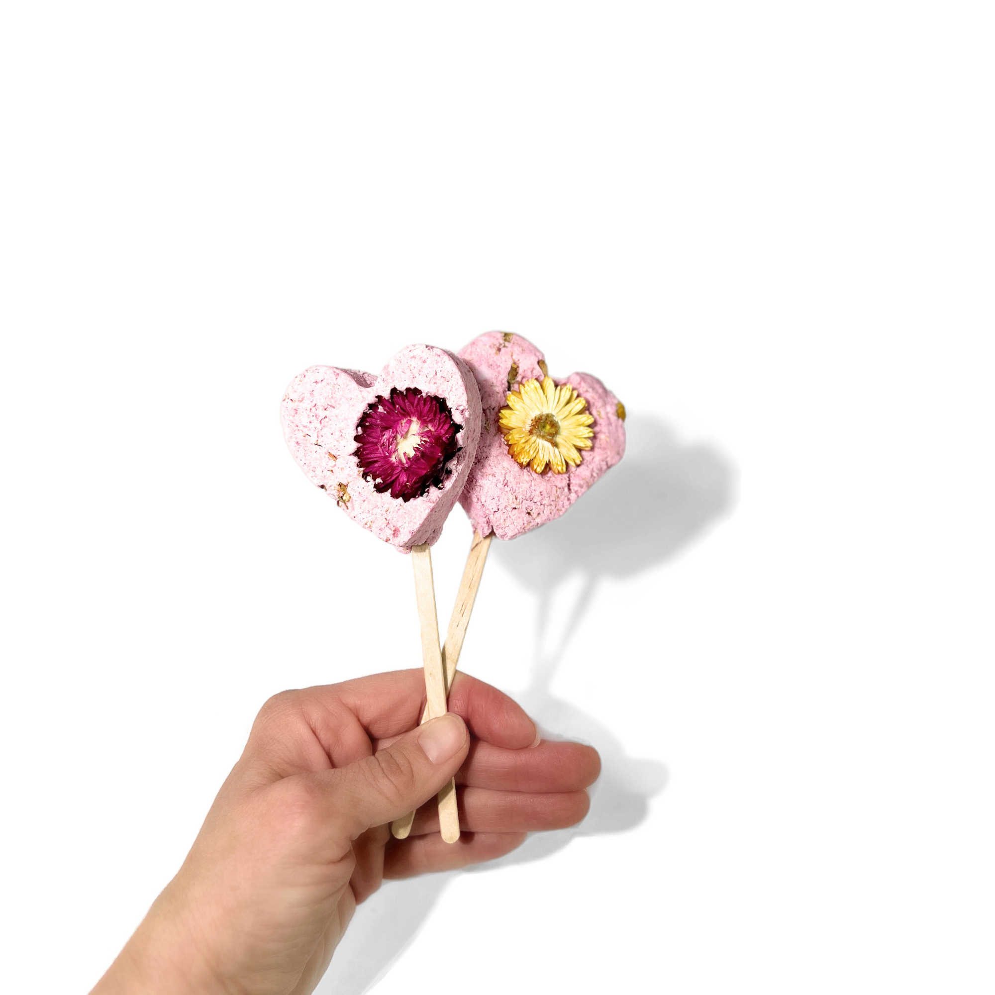 Hand holding two pink heart-shaped bath clay pops with yellow and magenta dried flowers, symbolizing love and care.
