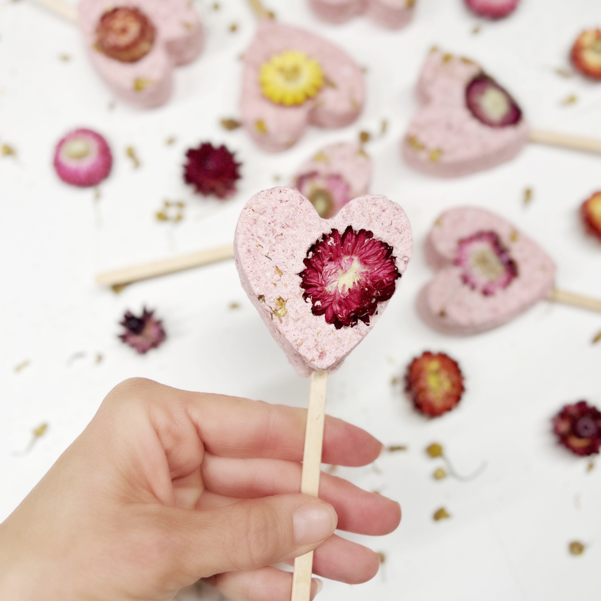 Hand holding a pink heart-shaped bath clay pop with a deep magenta dried flower in the center.