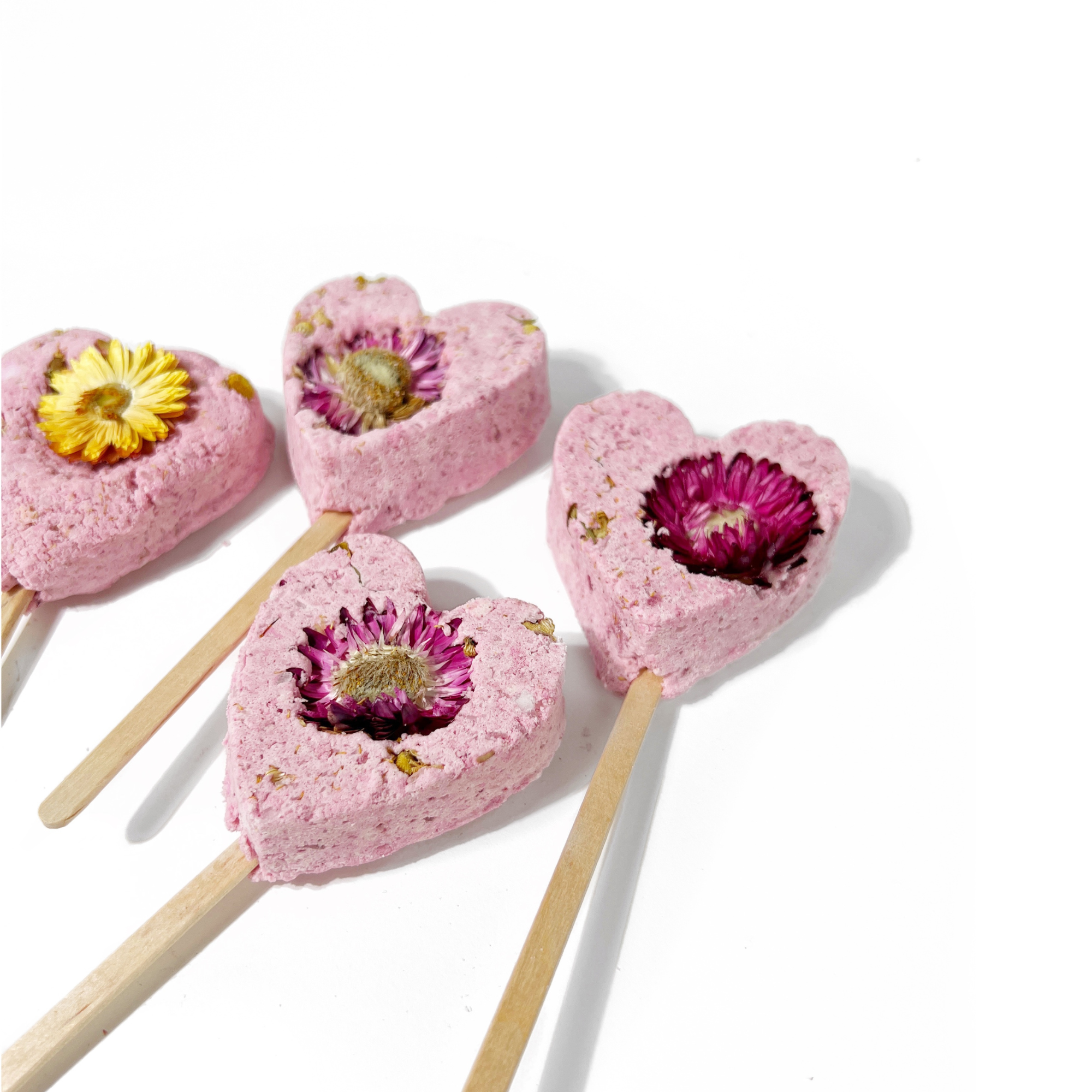 Close-up of pink heart-shaped bath clay pops on sticks, each with a colorful dried flower center.