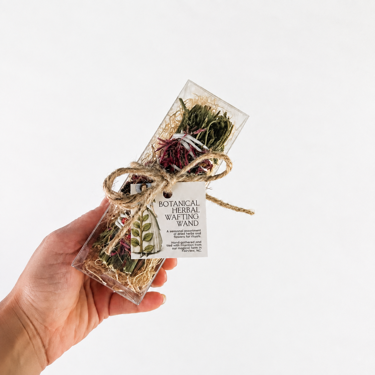 Hand holding a botanical herbal wafting wand in a clear gift box with twine and product tag
