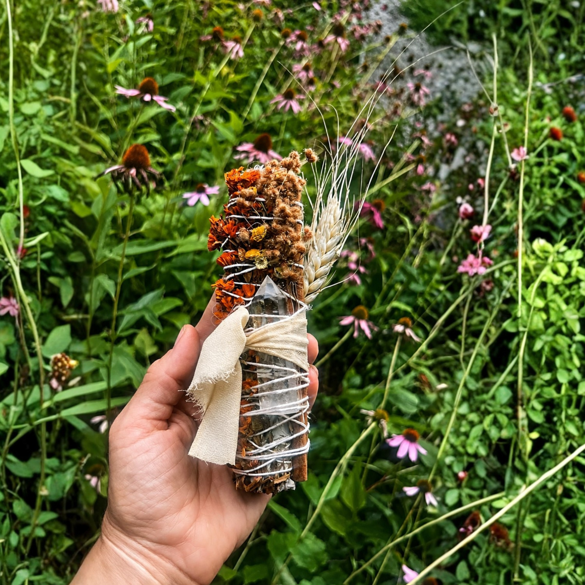 Hand holding a botanical smoke cleansing wand with dried flowers and herbs in a colorful garden setting