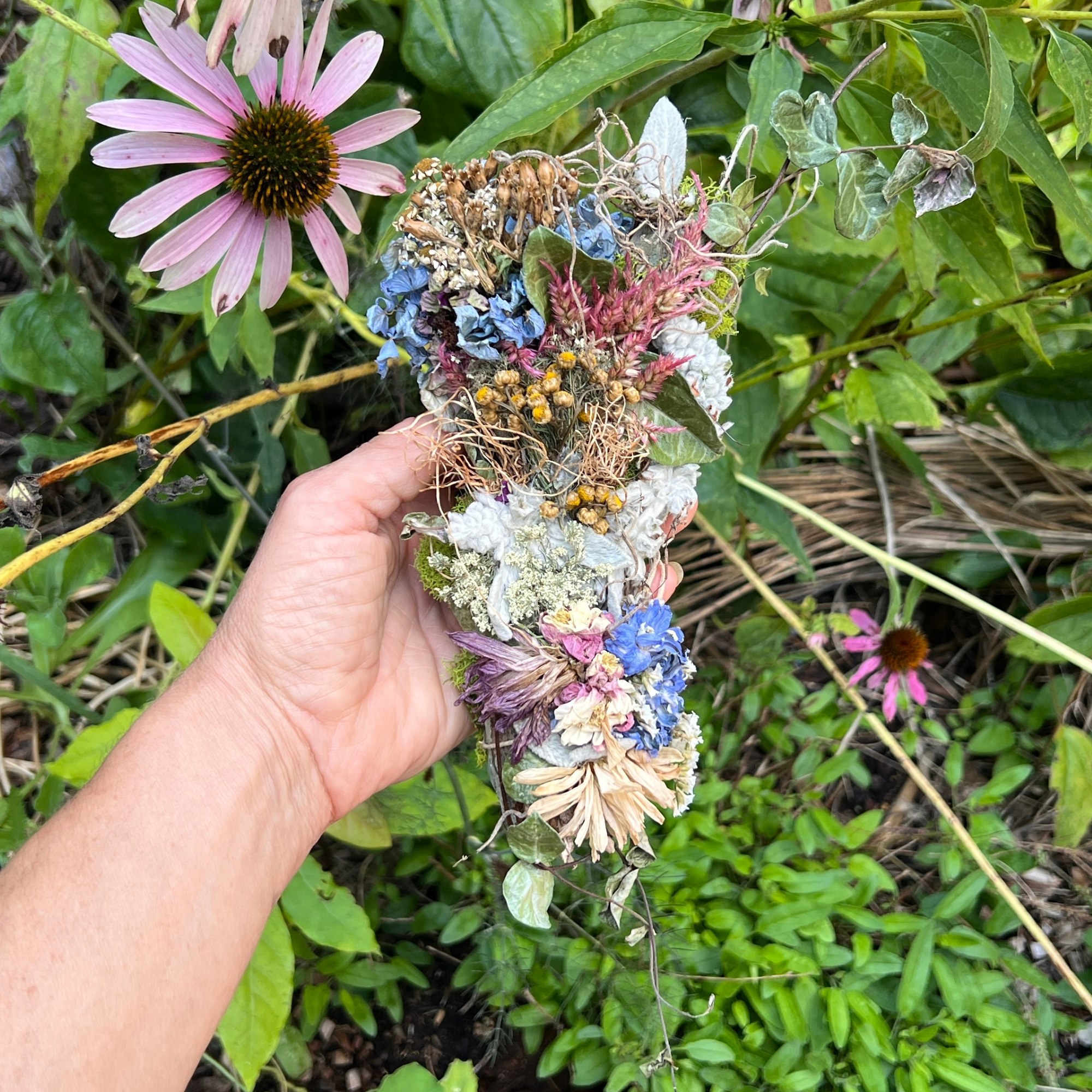 Colorful handmade botanical wall hanging displayed by hand among flowers and greenery in a garden