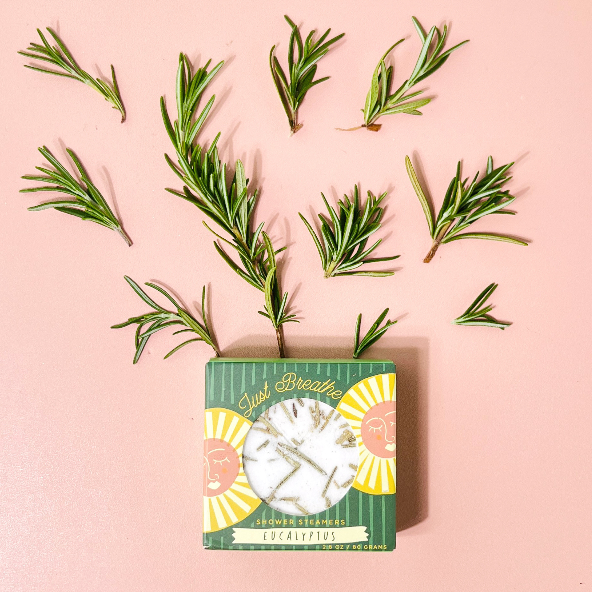 Eucalyptus shower steamer box surrounded by rosemary sprigs on pink background
