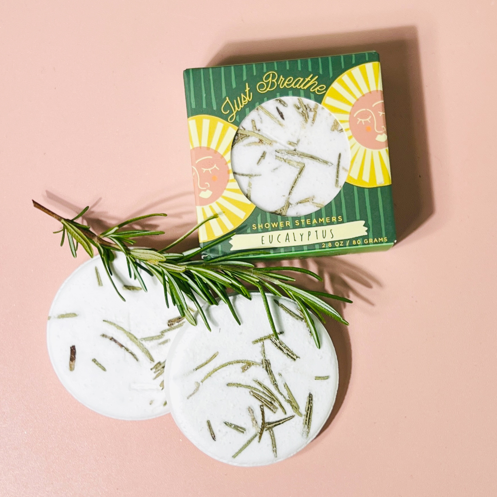 Two eucalyptus shower steamers with rosemary sprig beside open Just Breathe box on pink surface