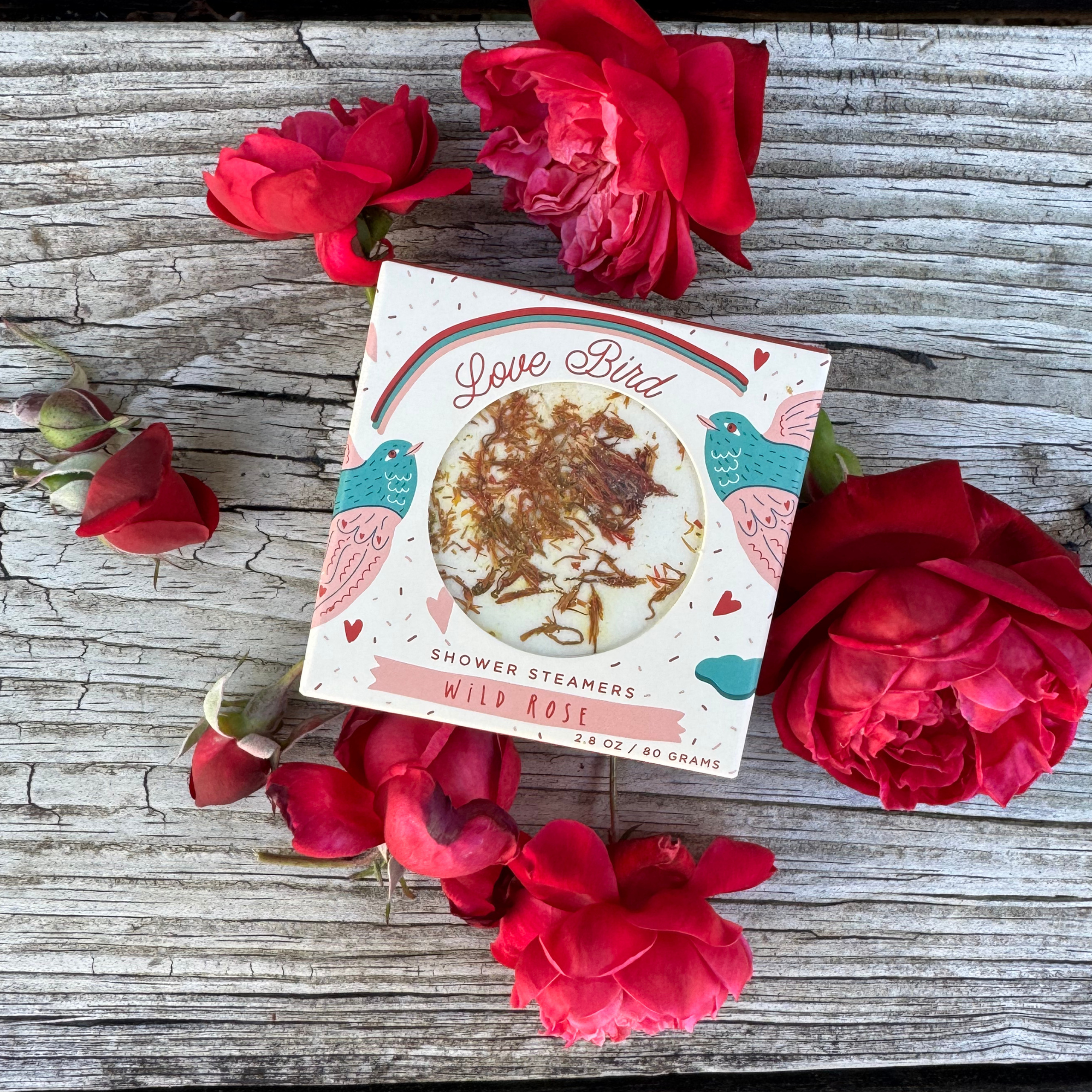 Love Bird wild rose shower steamer box surrounded by red rose blooms on wood table
