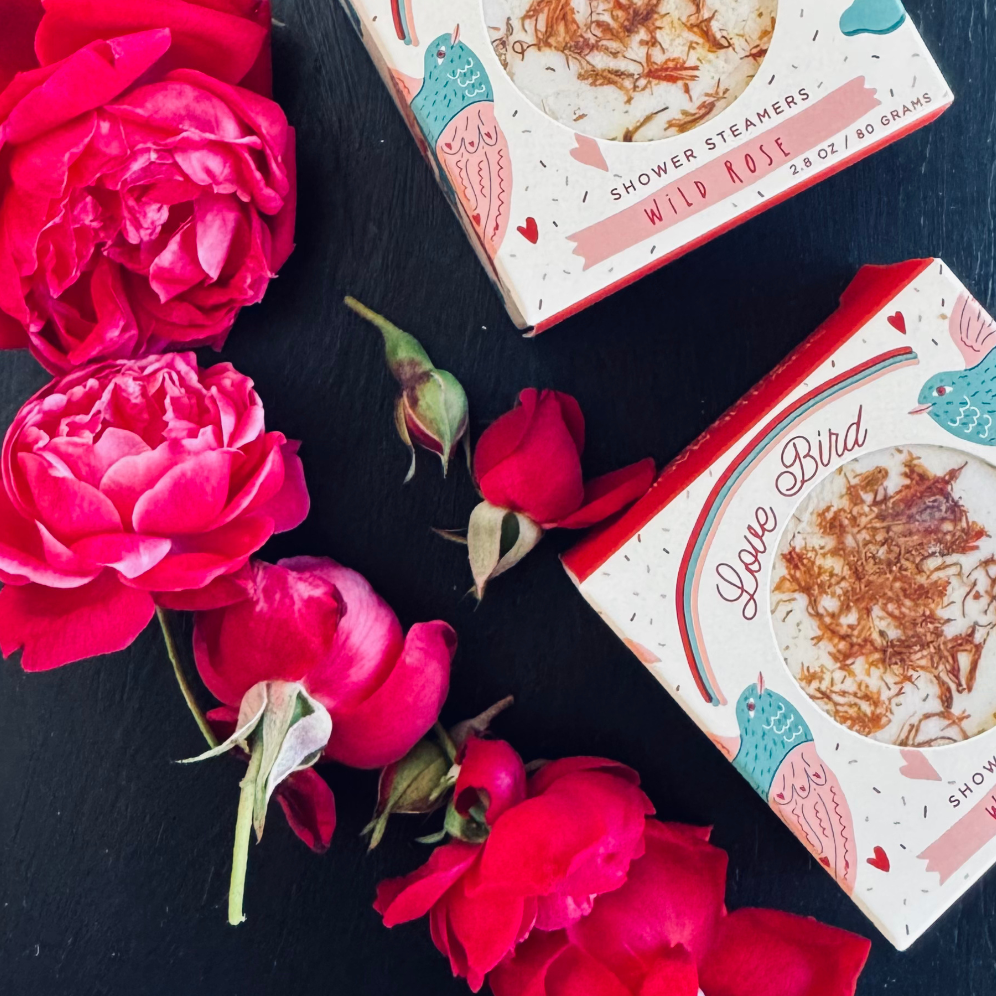 Love Bird wild rose shower steamers surrounded by fresh red roses on black background