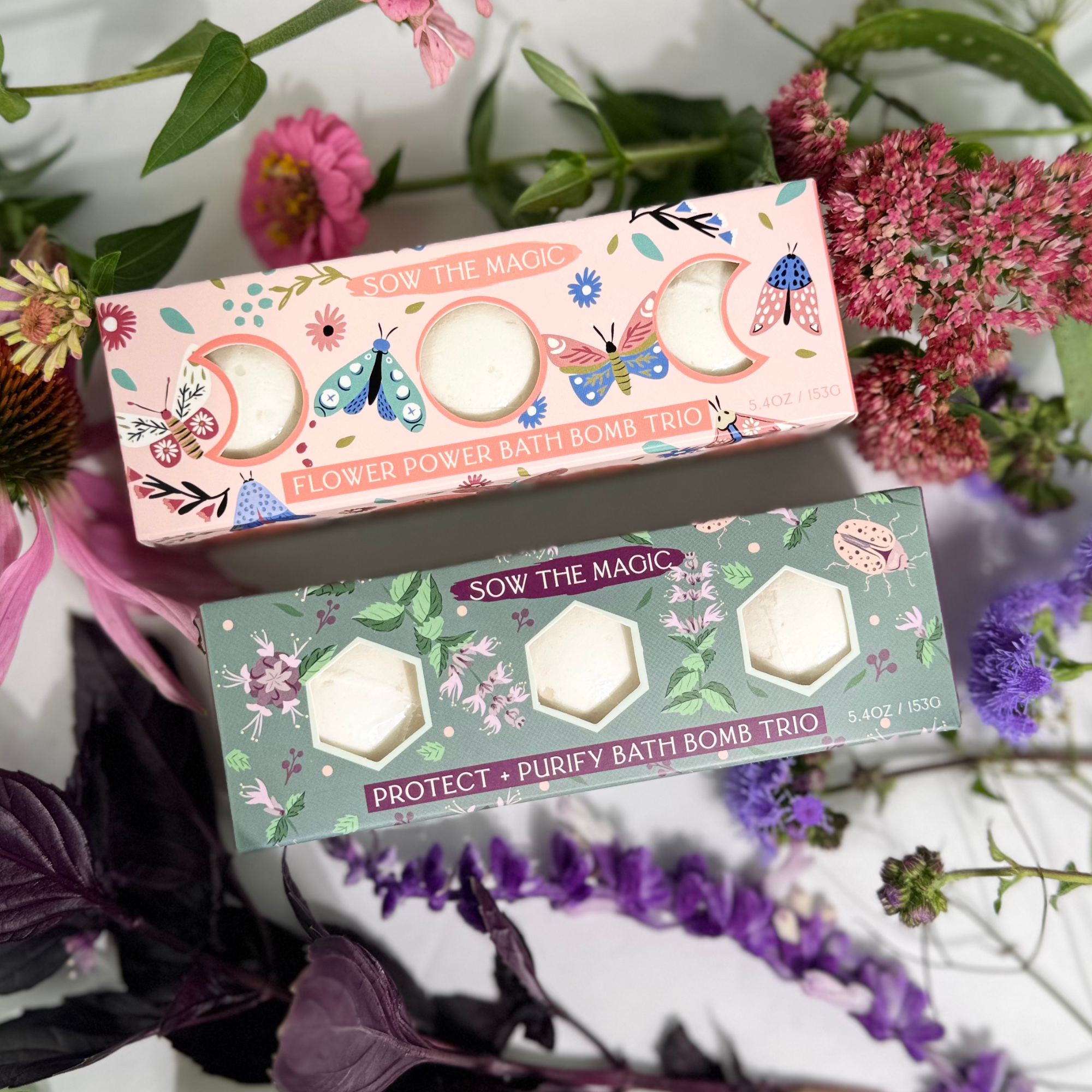 Protect + Purify and Flower Power bath bomb trios displayed with fresh herbs and pink and purple blooms