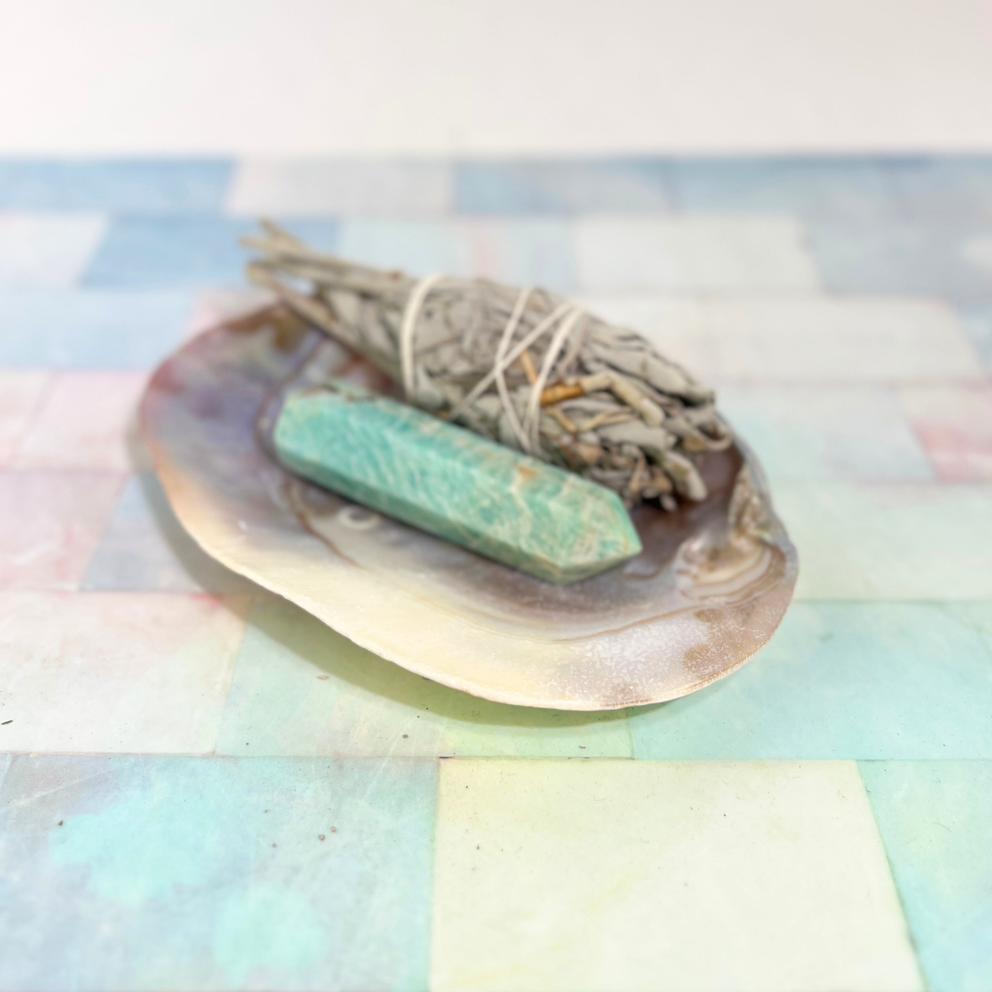 Pearl shell dish holding sage bundle and green crystal wand on soft pastel background