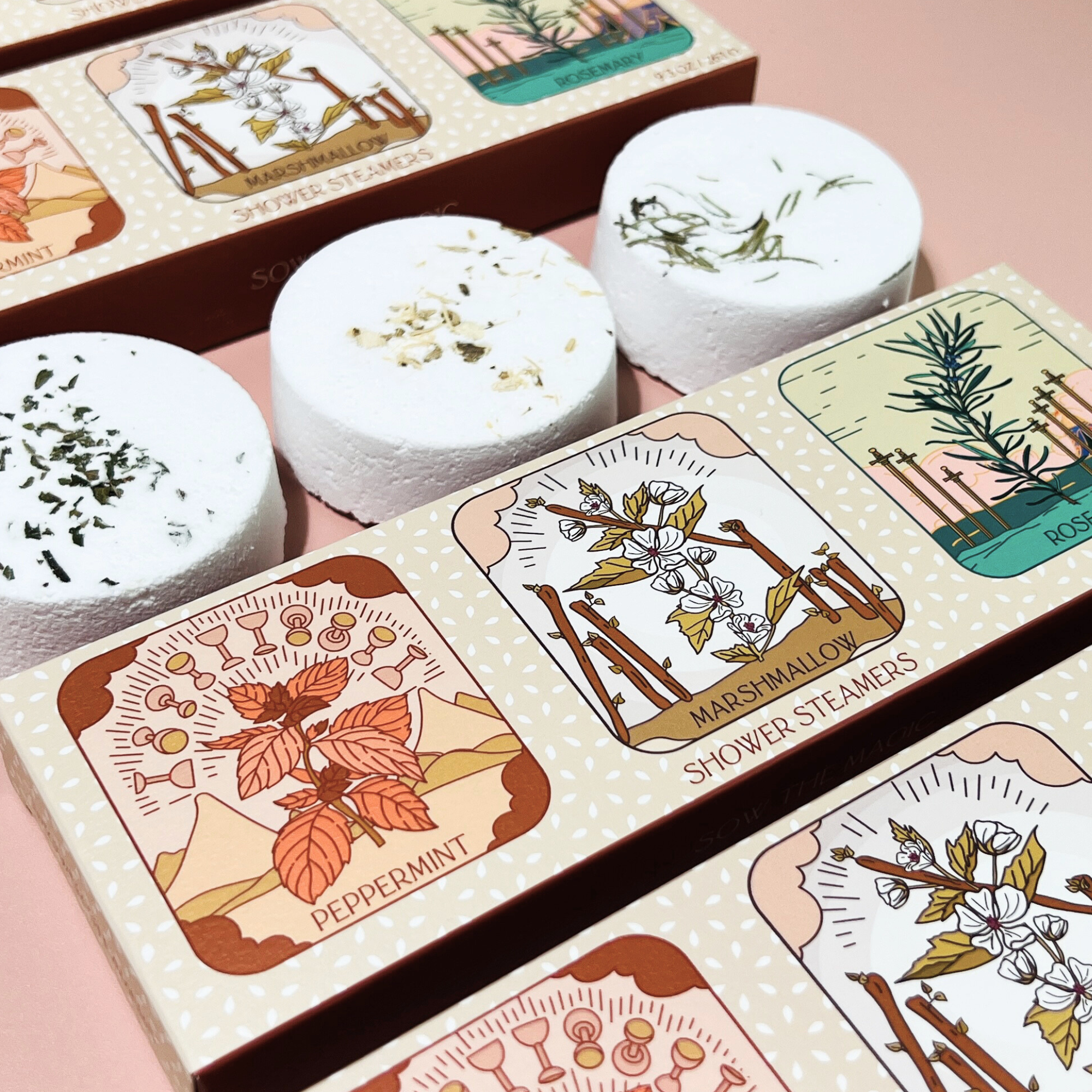 Close up of peppermint, marshmallow and rosemary shower steamers beside their illustrated tarot boxes