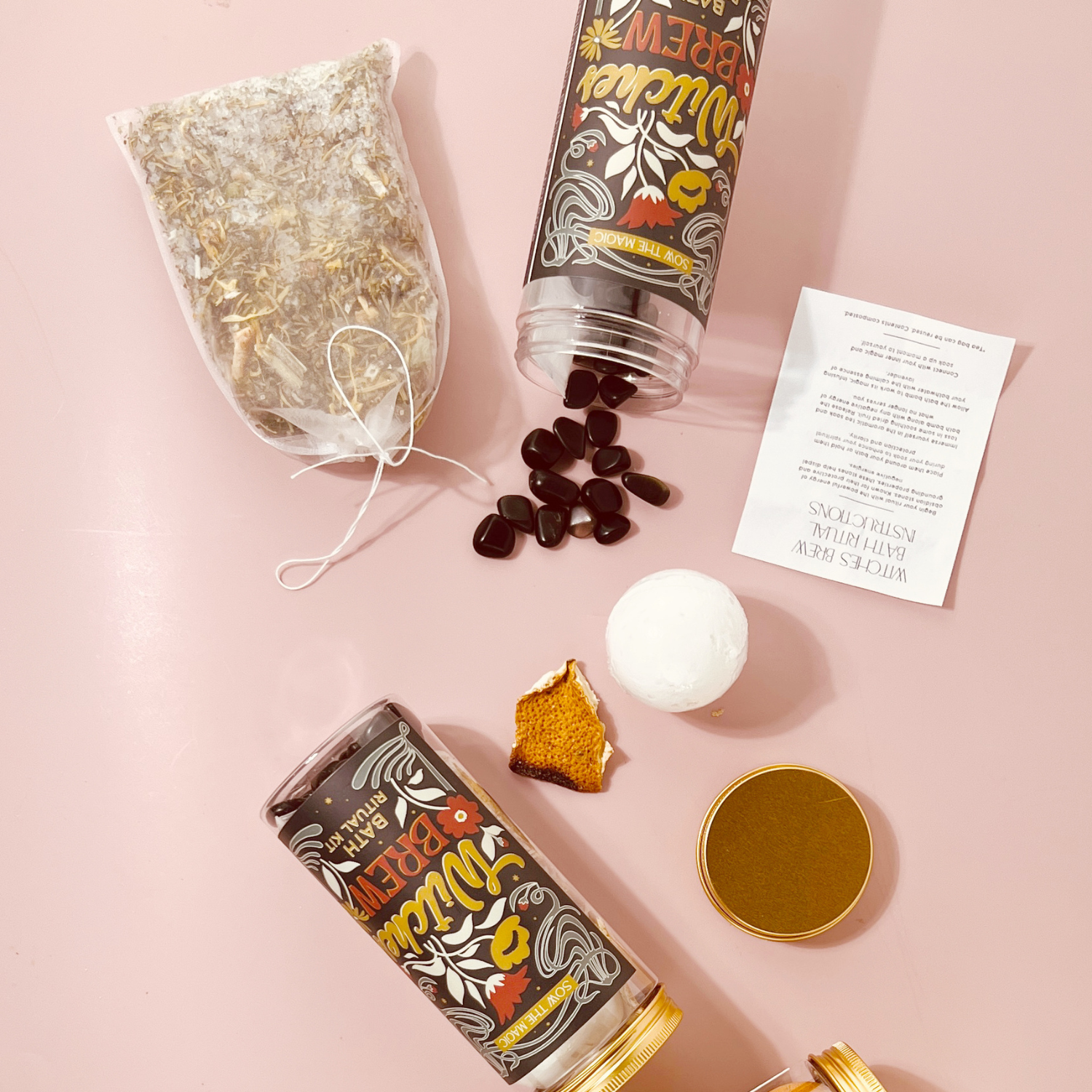 Witch’s Brew Bath Ritual Kit flat lay showing obsidian stones, herbal soak, bath bomb, and ritual instructions on pink background.