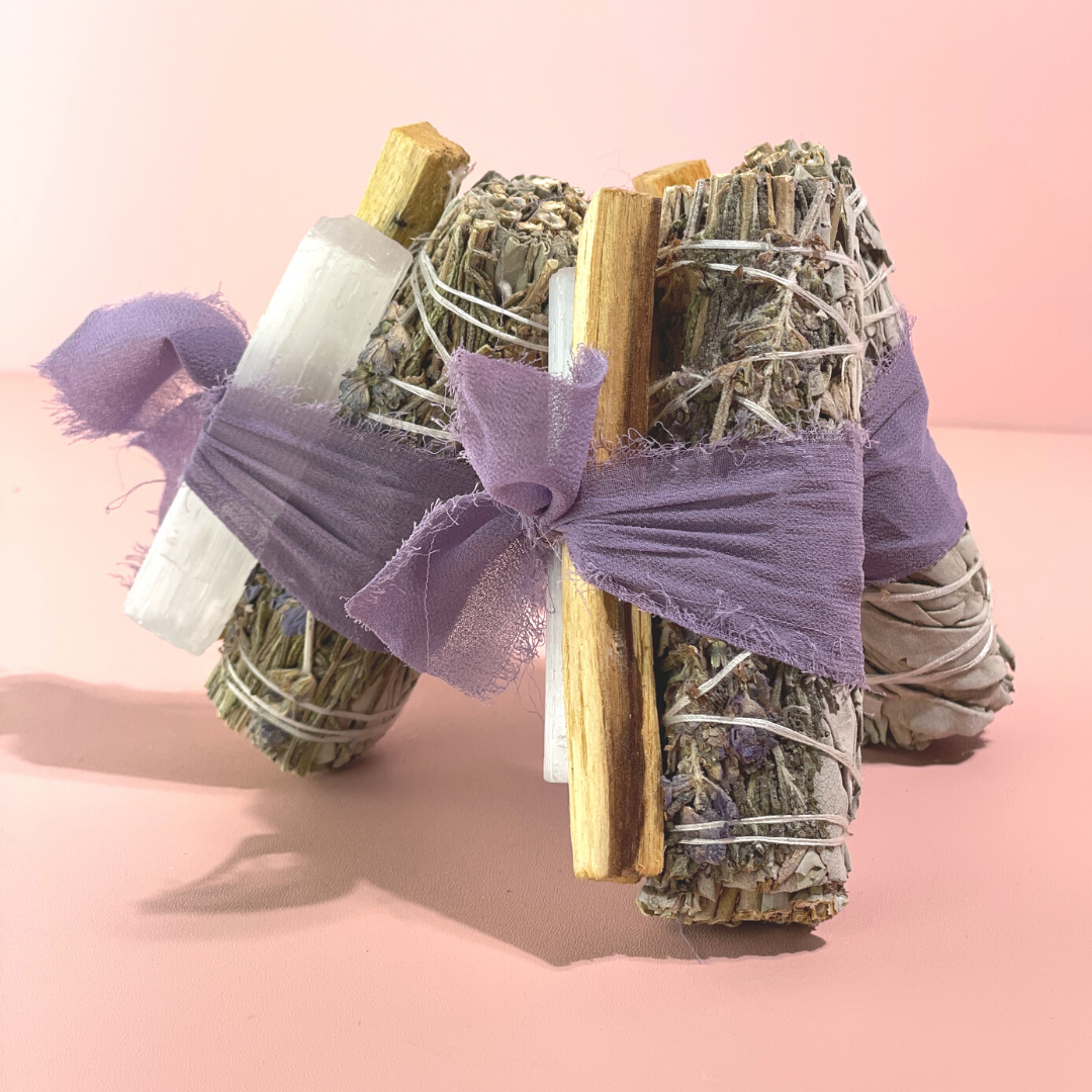 Flat lay of handcrafted White Sage Protection Ritual Bundles featuring White Sage, a lavender smudge stick, genuine Selenite cleansing wand, Palo Santo stick and a soft lavender ribbon arranged on a bright white background. Handmade spiritual cleansing gift set by Sow the Magic
