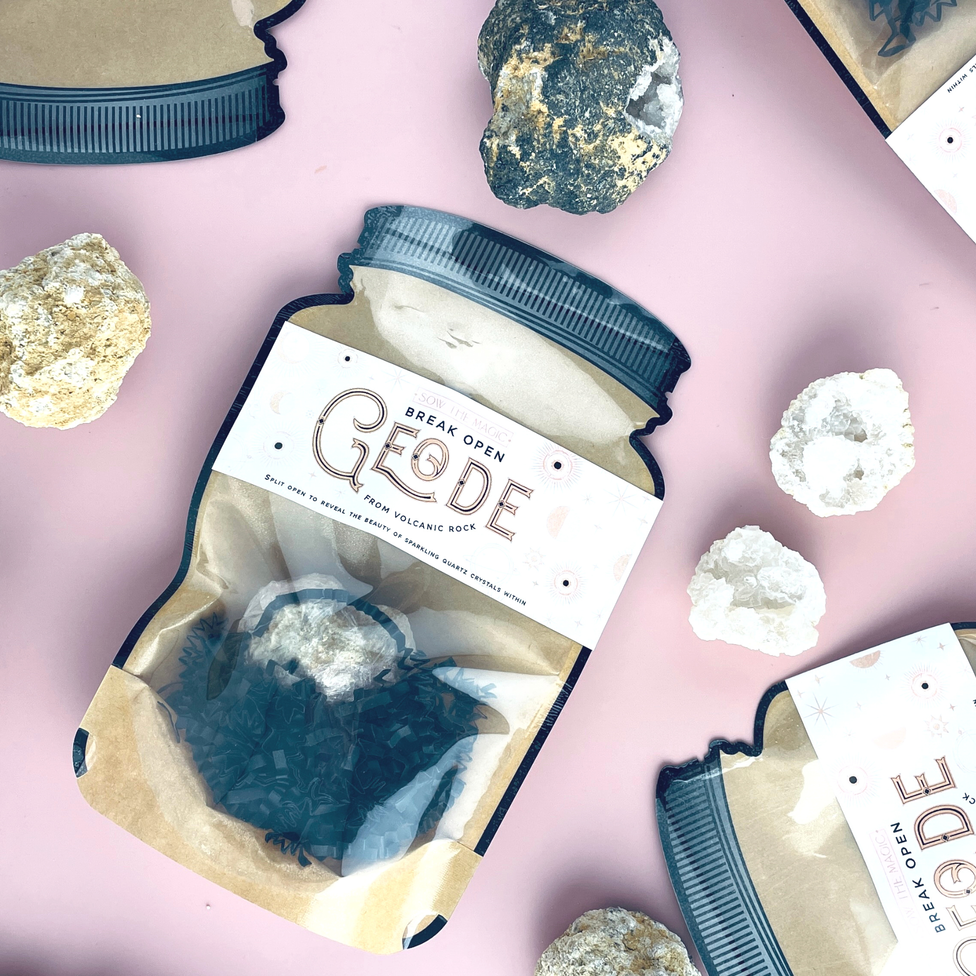 Flatlay of Break Open Geode Rock packaging with raw geodes and white quartz stones on pink surface