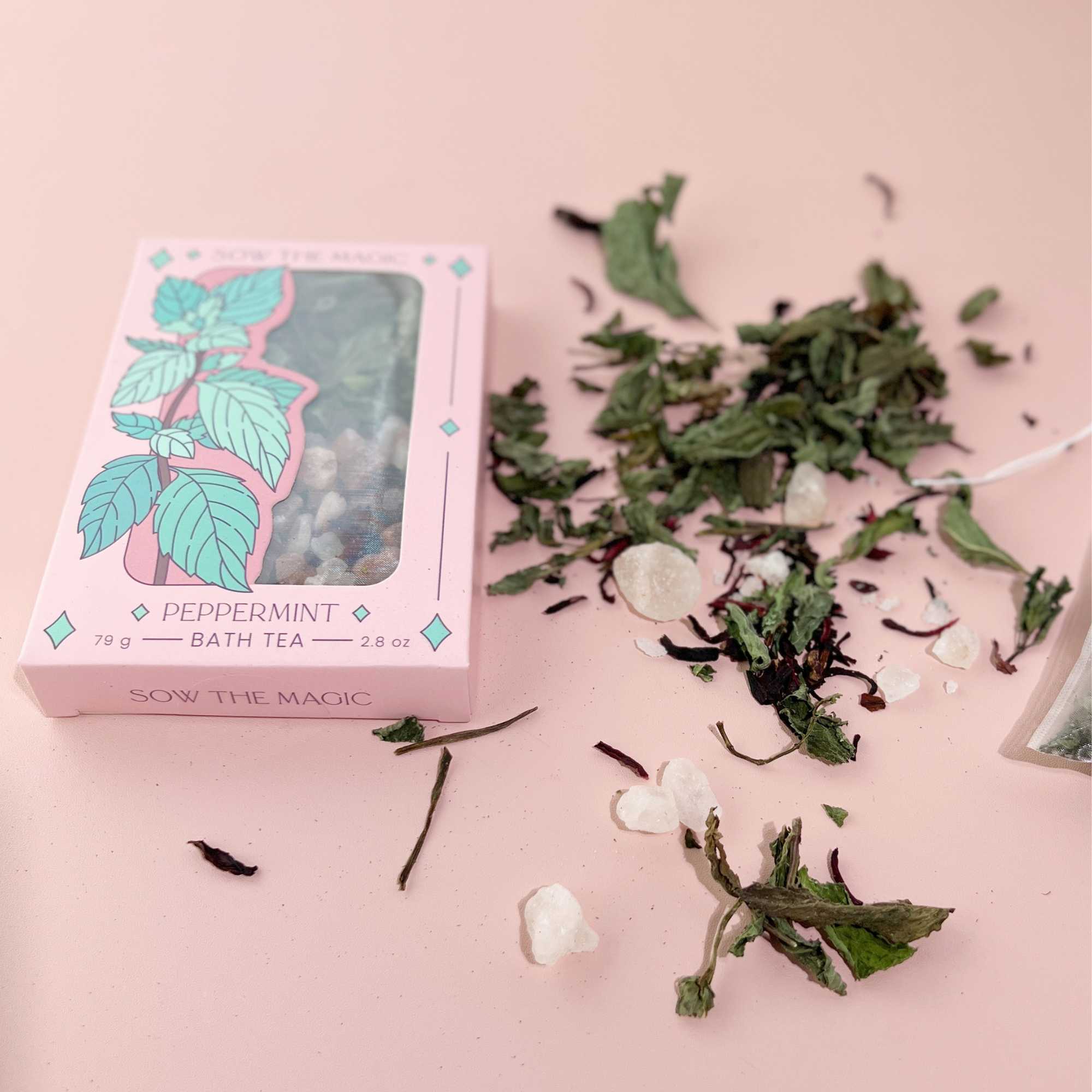 Peppermint Tarot Botanical Bath Tea Box surrounded by loose mint leaves, salts and botanicals on soft pink background.